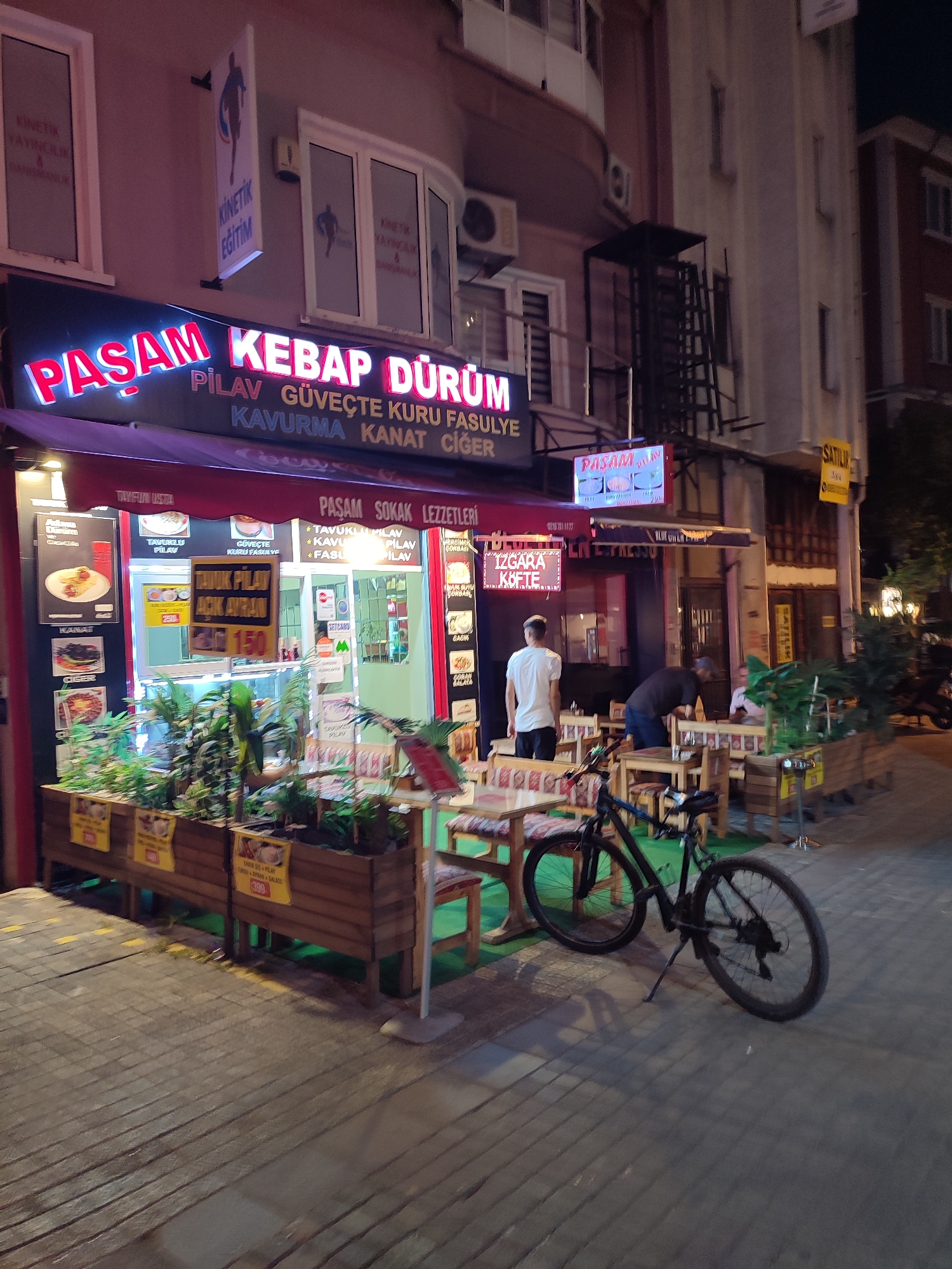 The image captures a street-level view of a restaurant facade and outdoor seating area in Kadıköy, Turkey. The restaurant, named "PASAM KEBAP DÜRÜM," features a prominent illuminated sign in red and blue neon, advertising various kebab dishes and "pilav" (rice). Below this main sign, smaller text lists items like "GÜVEÇTE KURU FASULYE" (dry beans in a casserole) and "KAVURMA KANAT CIGER" (roasted meat, wings, liver).
The establishment has a red awning and displays a menu with pictures of food items. Some of these appear to be "Tavuklu Pilav" (chicken pilaf) and "Güveçte Kuru Fasulye." Outdoor seating is arranged with wooden tables and benches, some of which are covered with patterned cushions. Plants in wooden planters are positioned in front of the seating area.
Two individuals are visible. One man, wearing a white t-shirt and dark shorts, stands near the entrance. Another man, in a dark t-shirt, is interacting with someone seated at one of the outdoor tables. A black bicycle is parked beside the outdoor seating area, leaning on its kickstand.
The overall environment suggests a commercial street in Kadıköy, likely in the evening or at night, given the illuminated signs and the ambient lighting. The buildings in the background are typical of urban architecture. Another sign, "SATILIK" (for sale), is visible on a building further down the street.