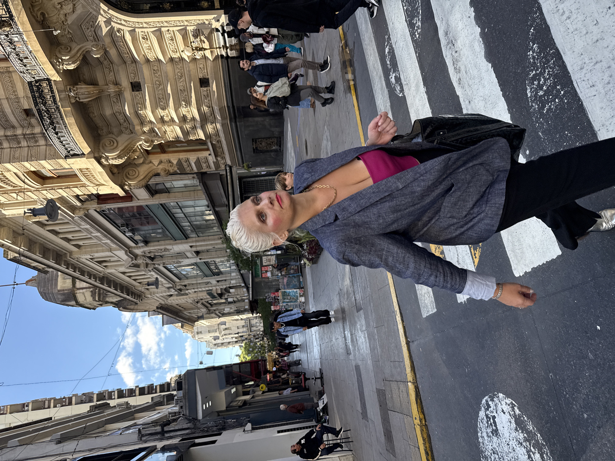A medium eye-level full shot features a fair-skinned, light-haired woman with red lipstick and pink blush, wearing a gray blazer and a fuchsia top, standing on a crosswalk in Buenos Aires, Argentina. She is facing towards the camera, while looking at the left of the frame. She is wearing black pants and has a black bag on her left shoulder. She is wearing a gold chain necklace.
A group of people, slightly out of focus, is behind the woman. Buildings with ornate facades line the street. The sky is blue with some white clouds. The overall lighting is bright and natural. The city is bustling with people.