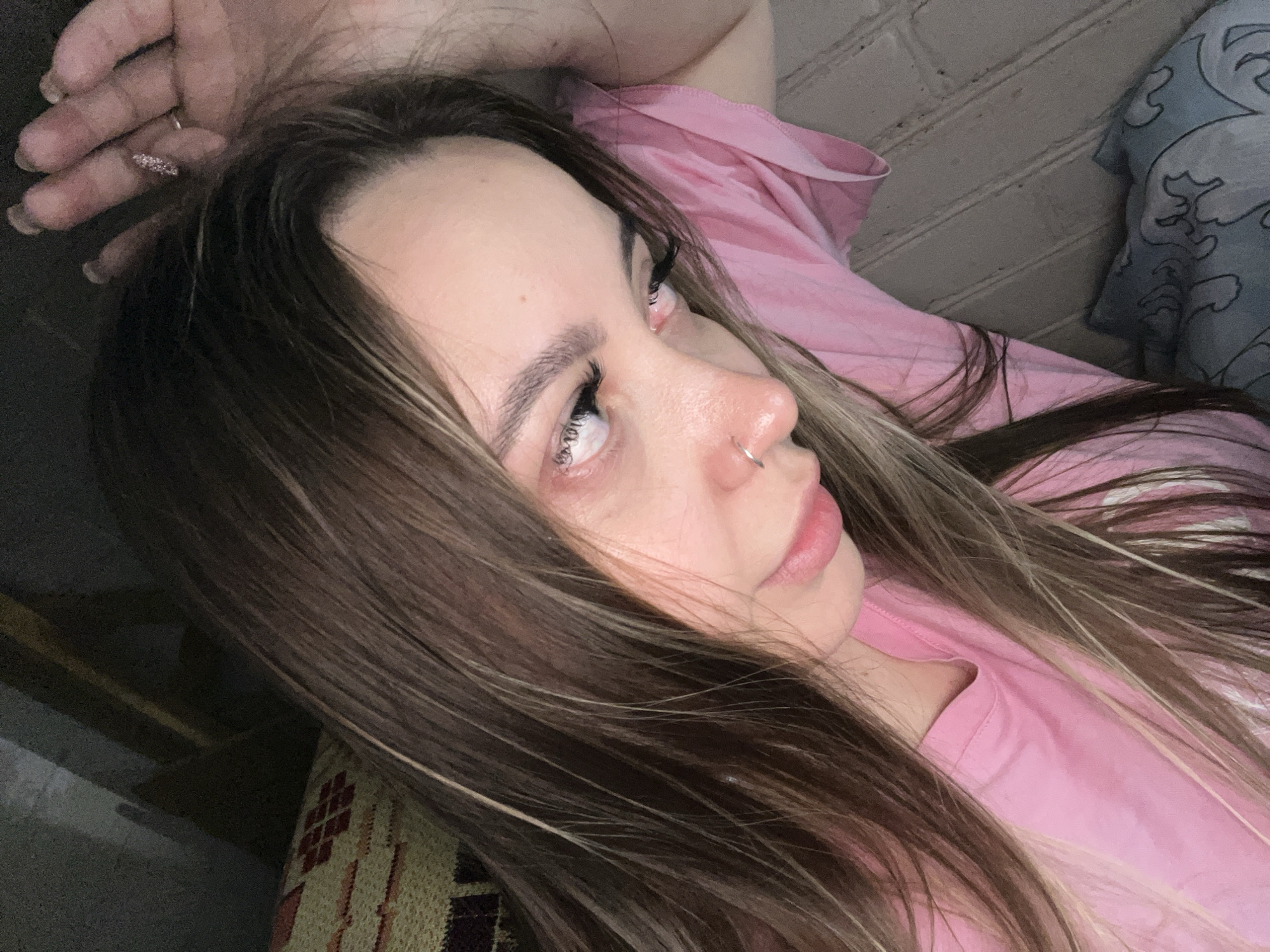 A woman with brown hair and a nose ring, wearing a pink shirt, is lying down. Her hand is raised above her head, and she appears to be looking upwards. The background includes a brick wall and part of a patterned pillow or cushion. There is no information or indication that this image was taken in Novopokrovskaya, Russia.
