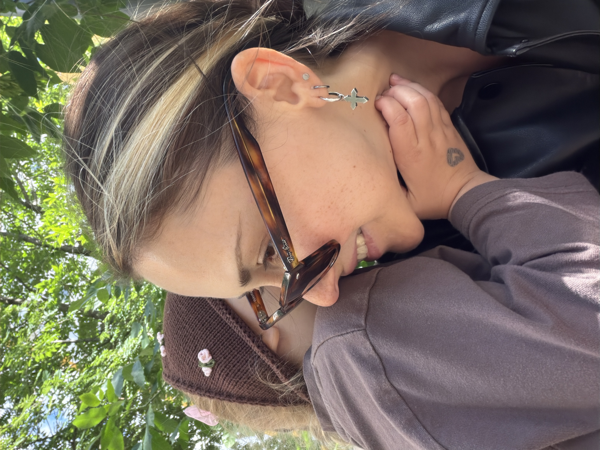 The image shows two people in close proximity outdoors. One person, in the foreground and facing right, is wearing sunglasses with tortoiseshell frames and a black leather jacket. They have a cross-shaped earring on their left ear and a small circular stud in the upper part of their ear. A small tattoo is visible on the back of their left hand. The second person, partially obscured and facing left, is wearing a brown knitted hat with small pink floral decorations and a light brown long-sleeved shirt. The background consists of green foliage and sunlight filtering through trees, suggesting a park or garden setting. There is no specific information within the image to indicate a location in Moscow, Russia.