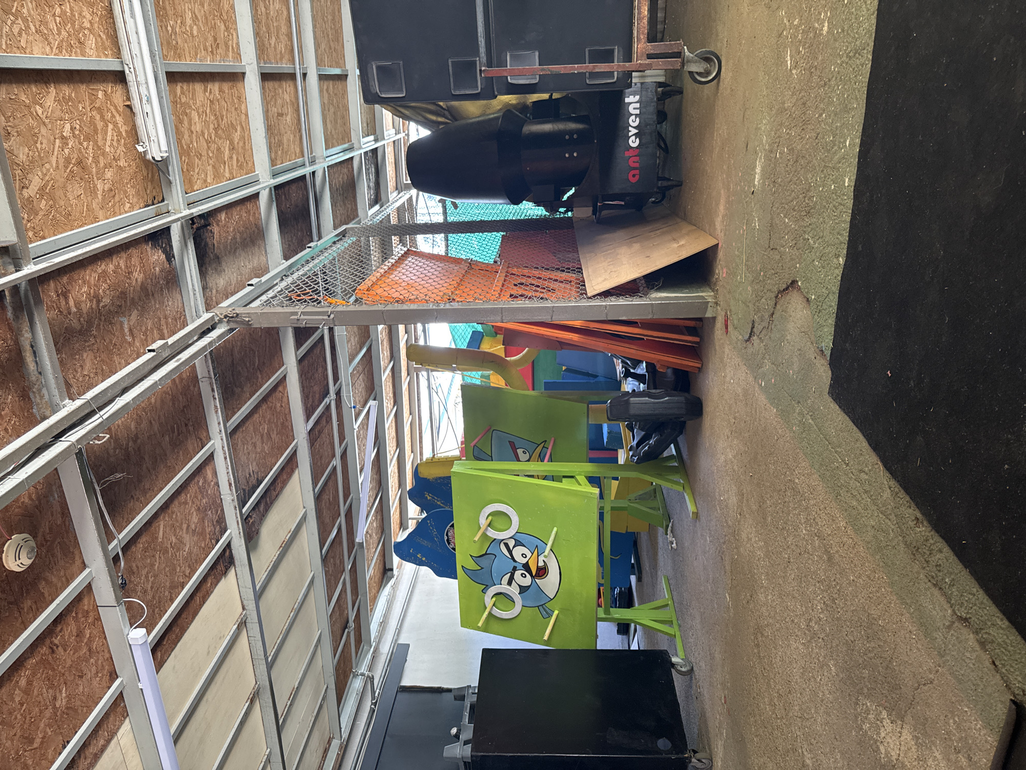 The image shows an indoor space, possibly a storage area or an events venue, with a concrete floor and a ceiling constructed of metal beams and wood panels. The space is cluttered with various objects, including a large black speaker stack on the right, a black cabinet with wheels on the left, and several colorful game-related structures.
Prominently featured in the center is a green board with a cartoon character, resembling an Angry Bird, painted on it. This board is part of a game setup with rings and yellow sticks. Behind it, other colorful structures, possibly parts of inflatable games or bouncy castles, are visible.
To the right, a large black conical object, which appears to be a fog machine, is situated on a cart with the "antevent" logo visible. A metal fence separates this area from other parts of the space. Orange planks and a piece of plywood are leaning against the fence.
The overall impression is one of an unorganized, utilitarian space filled with equipment likely used for events or entertainment. There are no people visible in the image. The lighting appears to be artificial, coming from overhead fluorescent lights.
Based on the provided location of Aksu, Turkey, this scene could be within a warehouse, an event hall, or an outdoor covered area adapted for events in that region.