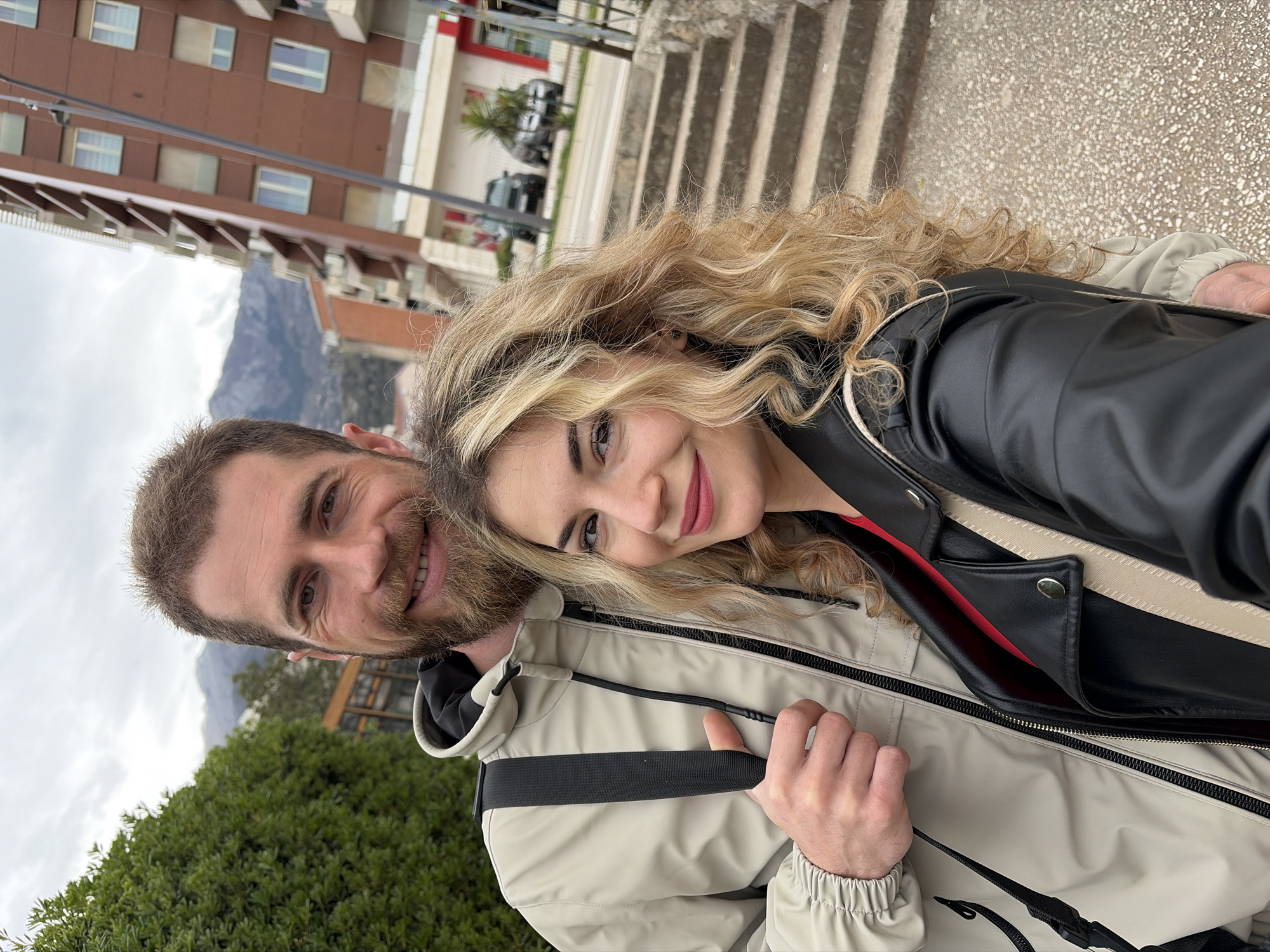 A selfie-style medium shot depicts a man and a woman in Budva, Montenegro. The man, on the left, has a brown beard and is smiling. The woman, on the right, has blonde curly hair and is also smiling. They are standing in front of a building, green bushes and a mountain range can be seen in the background. The building in the background is a modern structure with multiple windows. Concrete steps are near them. The sky is overcast. The woman wears a black leather jacket with a red shirt underneath. The man wears a beige jacket with black details.