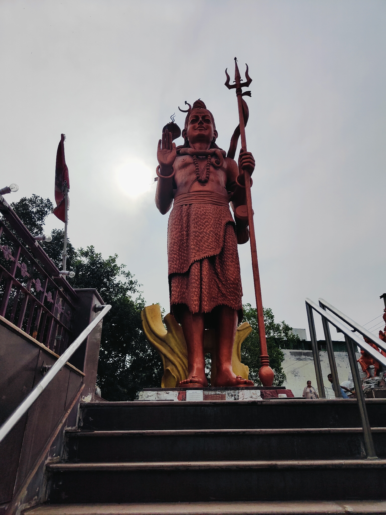 The image depicts a large, reddish-brown statue of the Hindu deity Shiva, holding a trident in his right hand and with his left hand raised in a blessing gesture. The statue is positioned in a public space, likely a temple or religious site, given the surrounding context. In the foreground, a dark set of stairs with metal railings leads upwards towards the statue. To the left of the statue, a red flag on a pole is visible, along with some trees and architectural elements of a building. The sky is overcast, with the sun peeking through from behind the statue, creating a lens flare effect. In the background, other smaller statues and structures can be seen, along with a few individuals walking around. This location is identified as being in Gurgaon, India.