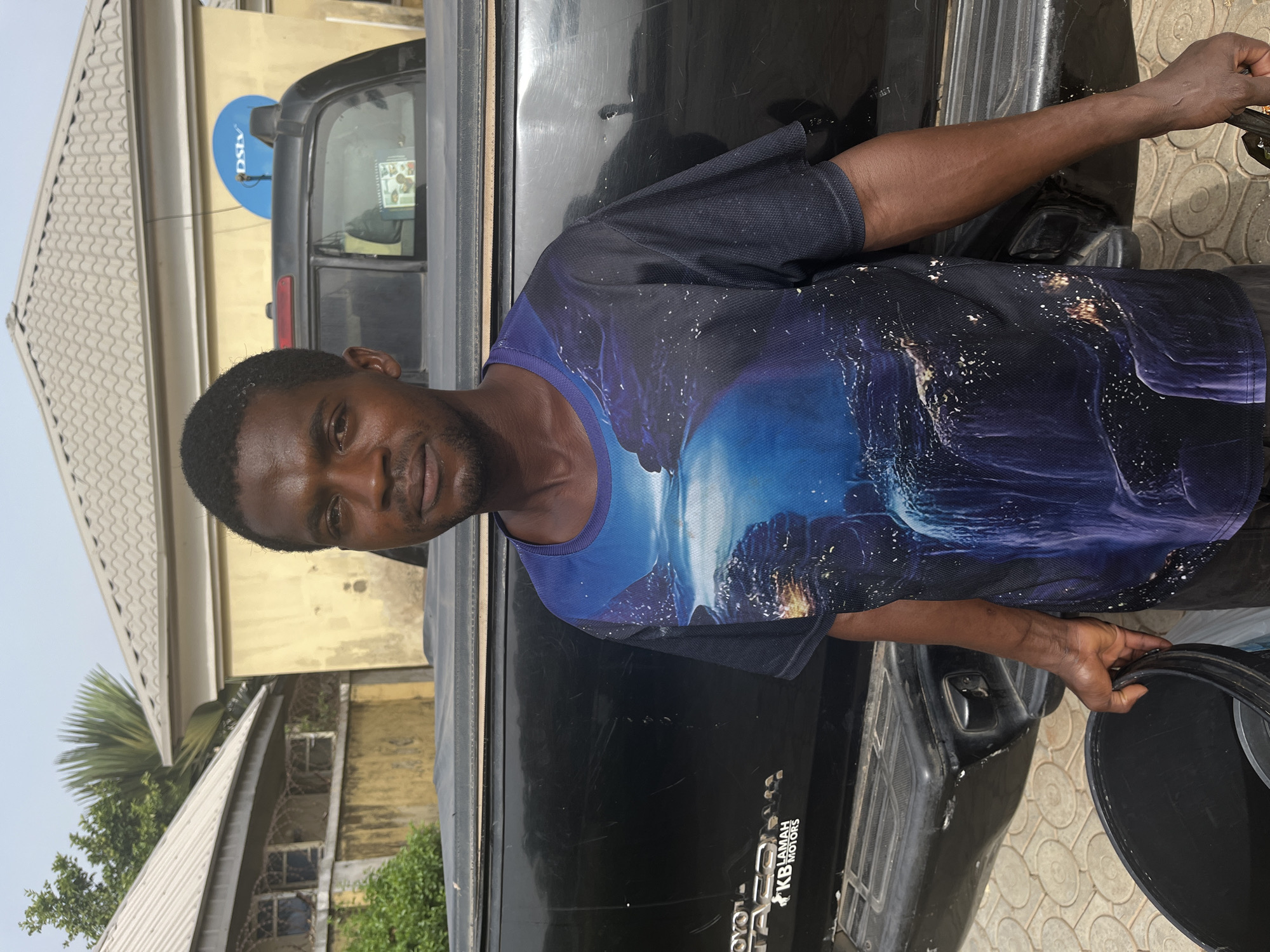 A medium shot, taken outdoors in daylight, shows a man standing beside a black Toyota pickup truck. The man has dark skin and short, dark hair. He is wearing a black t-shirt with a blue and white image of what appears to be an ocean scene. He looks at the camera. He is holding a black bucket. The pickup truck has a sticker that says "KB LAMAH MOTORS". In the background, there is a yellow building with a "DSTV" satellite dish on it, barbed wire, and trees. Given the sticker on the car, it is likely that the location is in Abuja, Nigeria, where KB Lamah Motors is a known entity.
