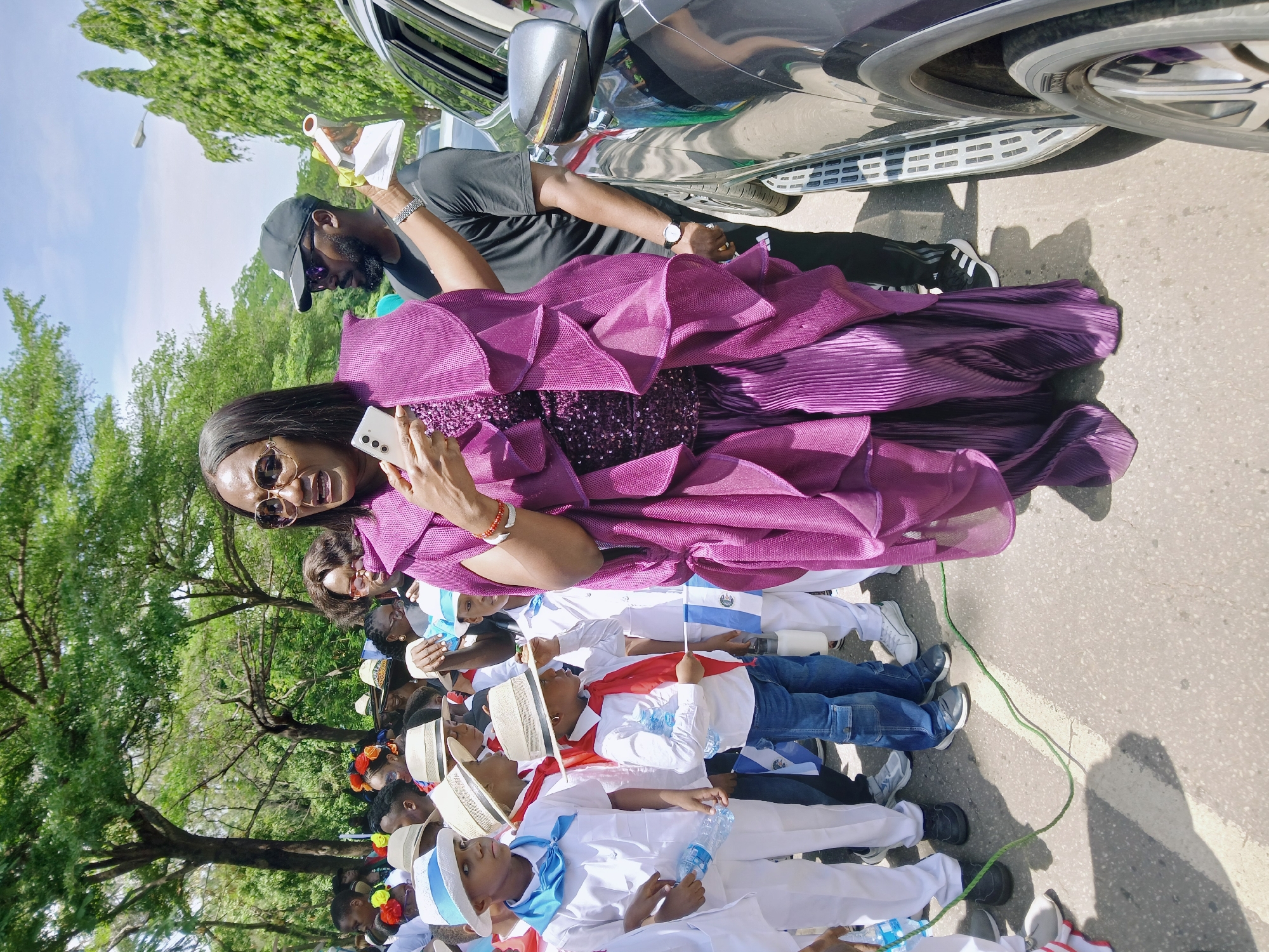 A medium, eye-level shot shows a woman in a purple gown taking a selfie with children lined up behind her in Abuja, Nigeria. The woman is fair-skinned, wearing oval sunglasses, and holding a white smartphone. She is positioned to the left of the frame, smiling at the camera. The gown is adorned with a sparkly sequin bodice and layers of ruffles at the top and pleated fabric on the skirt. A man in a black t-shirt and a baseball cap is standing behind her, holding a white container.
The children behind her are dressed in white shirts and pants with blue and white hats. They are holding blue and white flags and some hold bottles of water. They are connected to one another with a green rope on the floor.
A silver car is parked on the upper right of the frame, with its door open and a side step is visible. The scene is an outdoor, sunlit area with a tree line along the left side, suggesting a street or a park in Abuja, Nigeria. The road surface is visible.