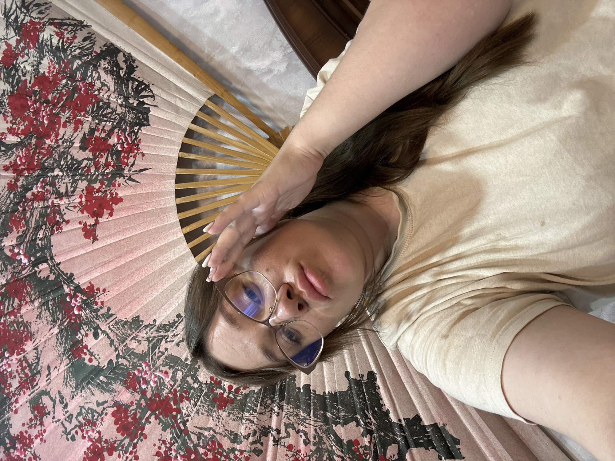 A woman with brown hair and glasses is holding an Asian-style folding fan with red and black floral designs on a beige background. She is wearing a beige T-shirt and her right hand is near her forehead. Part of what appears to be a dark-colored piece of furniture is visible in the background. Location information is not discernible from the image.