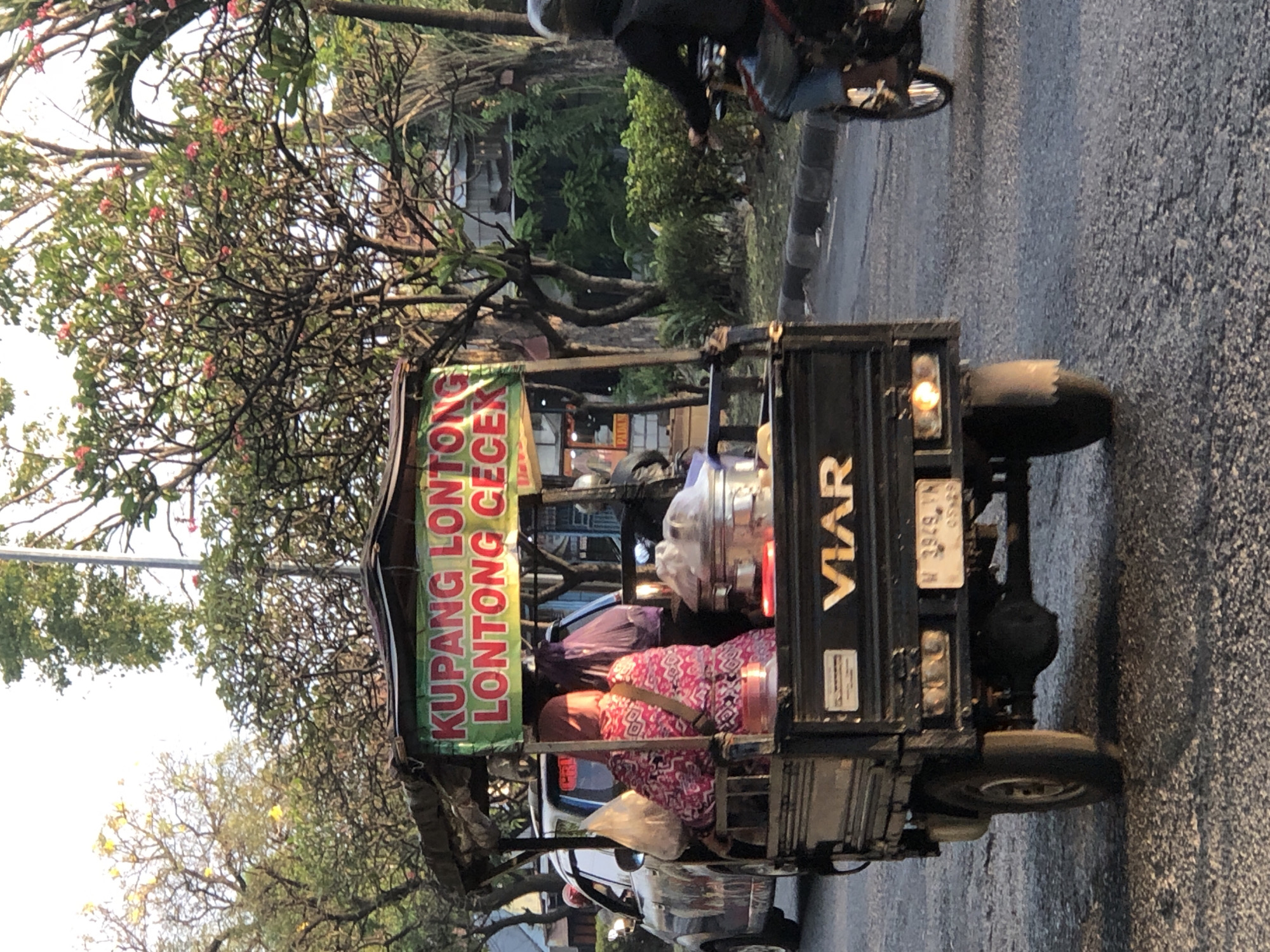 A vendor in a three-wheeled vehicle is driving down a street in Gelam, Indonesia. The vehicle has a sign that reads "KUPANG LONTONG LONTONG CECEK". A person wearing a pink and white patterned dress and a headscarf is seated in the back of the vehicle. The vehicle is carrying food items. A person on a motorcycle is driving behind the vendor vehicle. The street is paved and lined with trees. The sky is visible in the background, with some clouds.