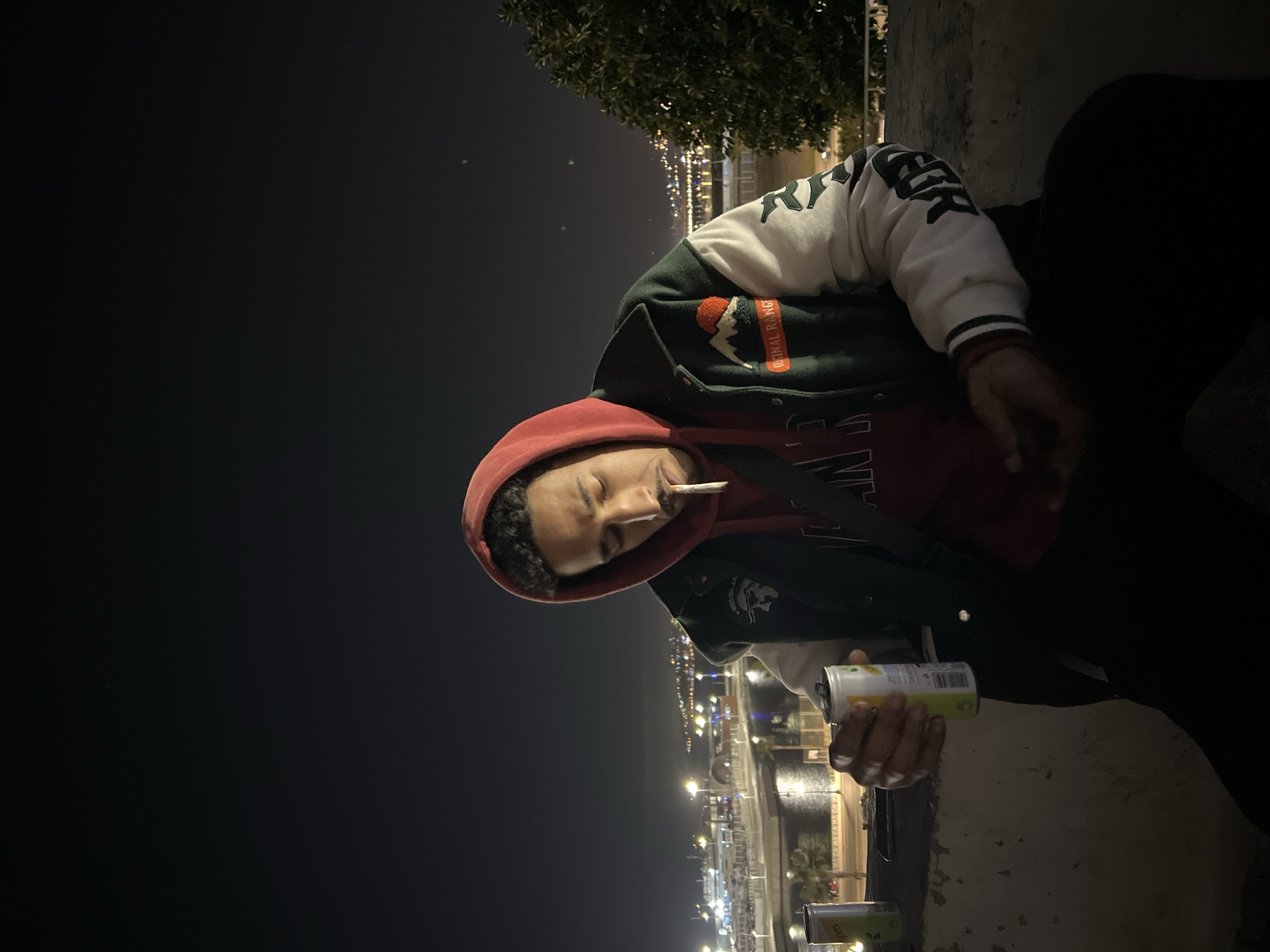 An individual is depicted at night, seated on a concrete ledge, overlooking the Port of Tanger Ville and the adjacent city in Tangier, Morocco. The subject is a male with dark curly hair, wearing a red hooded sweatshirt under a dark green and white varsity-style jacket with a "ORIGINAL RANG" patch featuring a red sun over white mountains on the left chest and dark script on the right sleeve. A cigarette is present in his mouth, pointed downwards. He holds a white and lime-green can, branded "LIPES," in his right hand. A second "LIPES" can and a dark smartphone are on the ledge to his left. In the background, city lights illuminate buildings, roads, and the port area where multiple large ferries are visible docked on the water. A section of foliage is visible on the right side of the frame.
