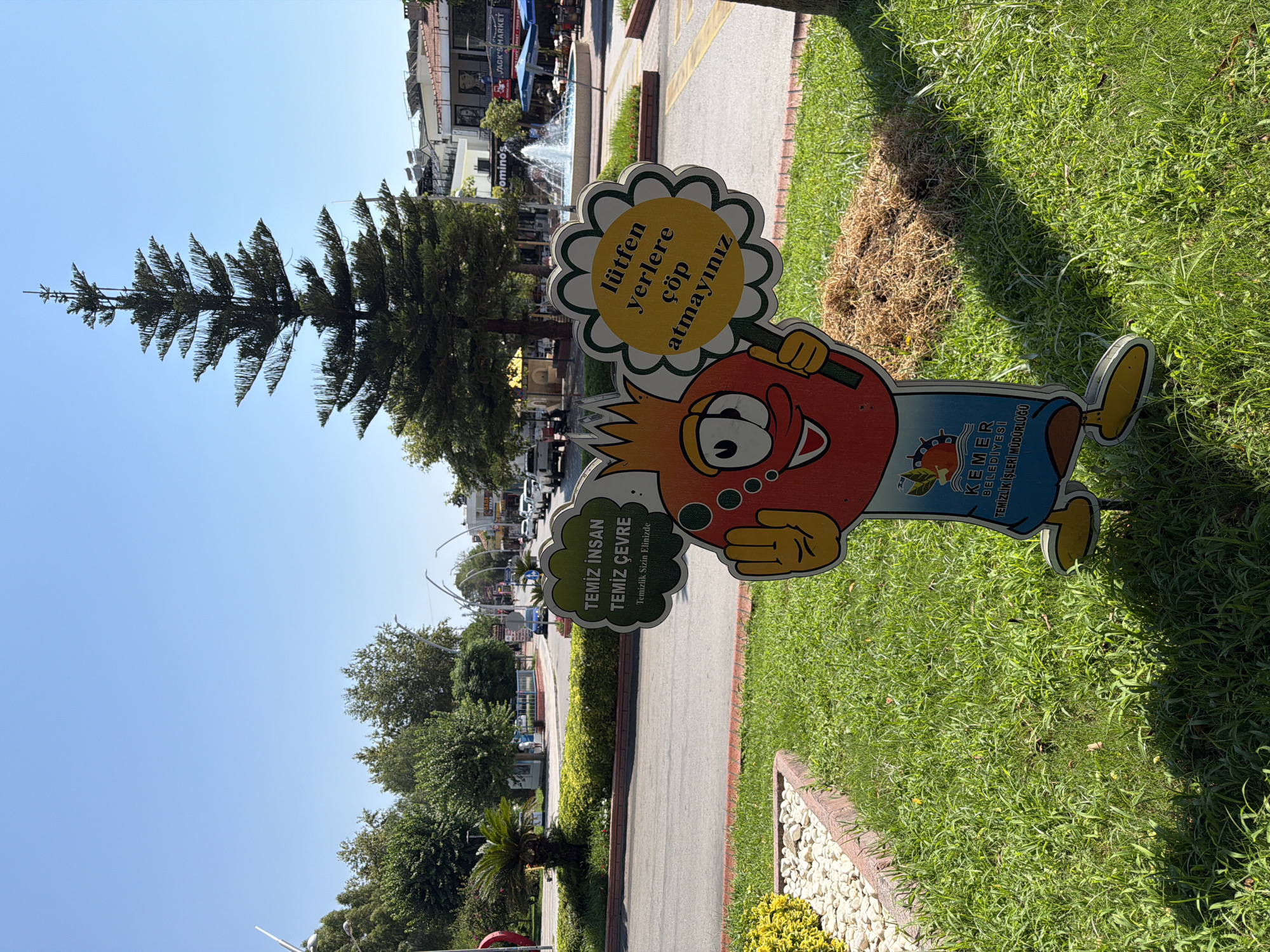 This is a medium shot, eye-level, outdoor photograph taken on a sunny day in Kemer, Turkey. The main subject is a cartoon character cutout with a pomegranate-shaped head and a blue body, standing on a grassy area. The character is holding a sign in the shape of a flower with yellow text and a green border, which reads "lütfen yerlere çöp atmayınız" (please do not litter). To the left of the character, there is another sign in a cloud shape, with green text reading "TEMİZ İNSAN TEMİZ ÇEVRE" (Clean Human Clean Environment) and smaller text below it "Temizlik Sizin Elinizde" (Cleaning is in Your Hands). The character cutout itself has a sticker at the bottom with text that includes "KEMER BELEDİYESİ" (Kemer Municipality) and "TEMİZLİK İŞLERİ MÜDÜRLÜĞÜ" (Directorate of Cleaning Affairs).
Behind the character, a street is visible with parked cars, a fountain, and buildings. The architecture of the buildings suggests a commercial area with shops and restaurants. A tall, conical evergreen tree stands prominently behind the fountain. The foreground is dominated by green grass and a decorative border with white stones. The sky is clear and blue. The overall scene depicts a public space in Kemer aiming to promote cleanliness and environmental awareness.