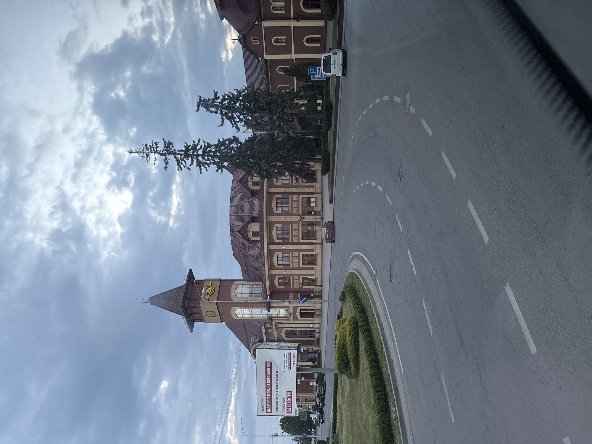 This is an eye-level, medium shot of the Uzhhorod railway station in Uzhhorod, Ukraine, under a cloudy sky. The station's main building, constructed of red brick with brown tiled roofs, features a prominent clock tower with a spire. To the left of the building is a large billboard advertising employment opportunities. In front of the station, a grey asphalt road curves to the right, with white dashed lane markings. Several cars are visible on the road and parked near the station. Two tall evergreen trees stand between the station and the road. The environment appears to be an urban setting, possibly in the early morning or late afternoon due to the lighting conditions.