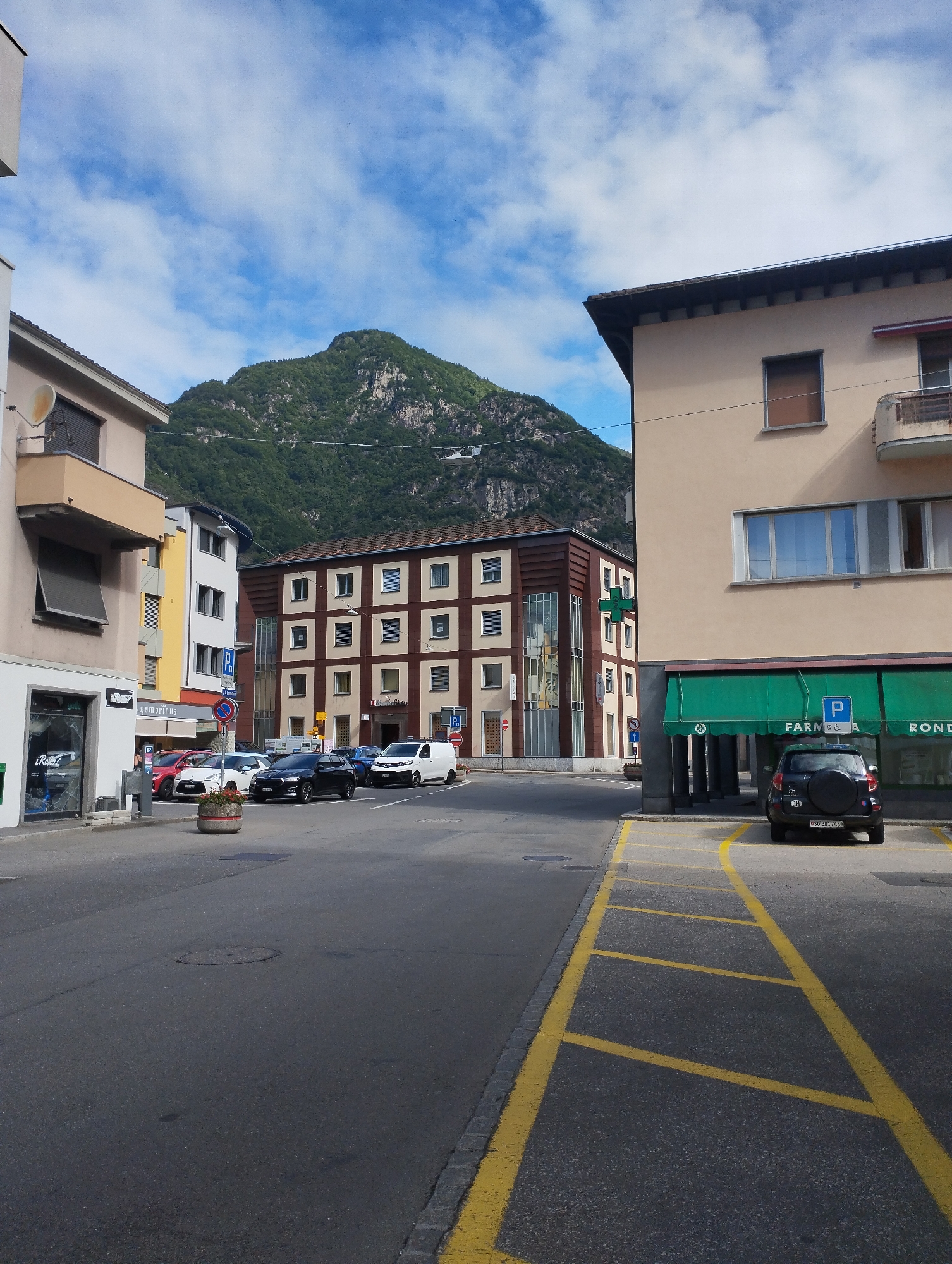 The image depicts a street scene in an urban environment. In the foreground, a street with marked parking spaces is visible, with yellow lines delineating the parking bays. A dark-colored SUV is parked in one of these bays. To the right, a building with a green awning and the word "FARMACIA" is present, indicating a pharmacy. Behind the pharmacy, another building with a beige facade and multiple windows is visible, with a balcony on the upper floor. To the left, a light-colored building with a multi-story design is present. In the middle ground, several cars are parked on the street, including a white van and a black sedan. Further in the background, a multi-story building with a distinctive architectural style featuring a grid of windows is located. Towering over the buildings and the town is a large, tree-covered mountain. The sky is a clear blue with scattered clouds. The scene appears to be a daytime shot, under good weather conditions.