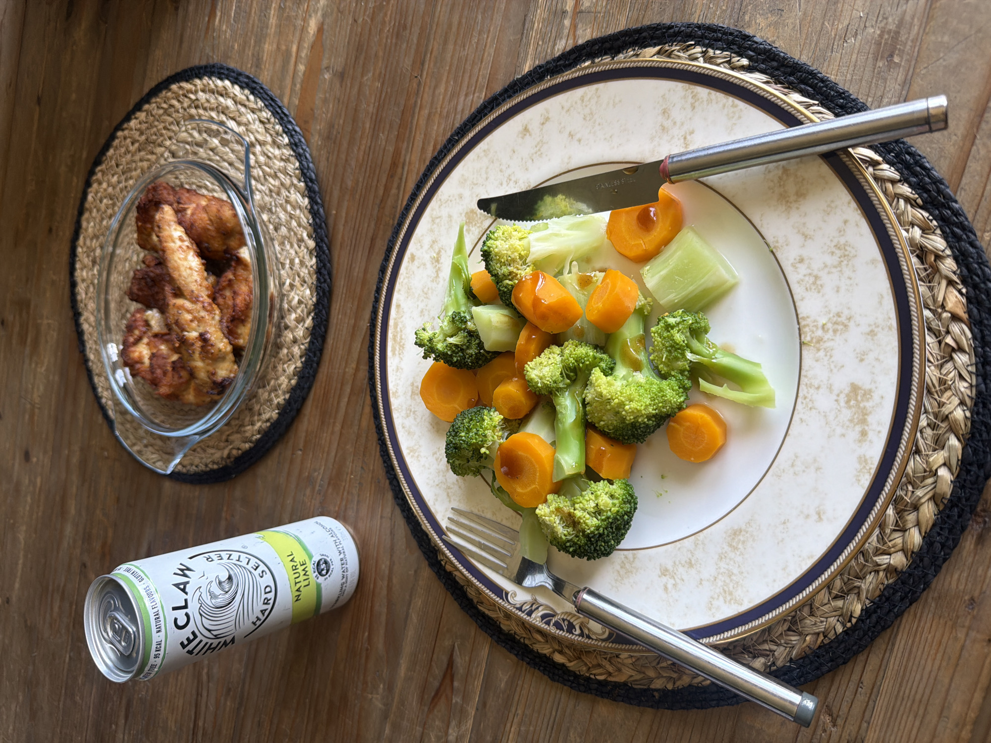 The image shows a meal laid out on a wooden table. In the foreground, a white plate with a decorative rim holds a portion of steamed broccoli and carrots. A fork is placed to the left of the plate, and a knife to the right. Behind the plate, a can of White Claw Hard Seltzer (Natural Lime flavor) is leaning against the edge of the table. To the right of the can, a glass bowl contains several pieces of what appear to be fried chicken. The entire scene is set on a textured wooden surface, with woven placemats under the plate and bowl. The lighting suggests an indoor setting, possibly a home dining area.