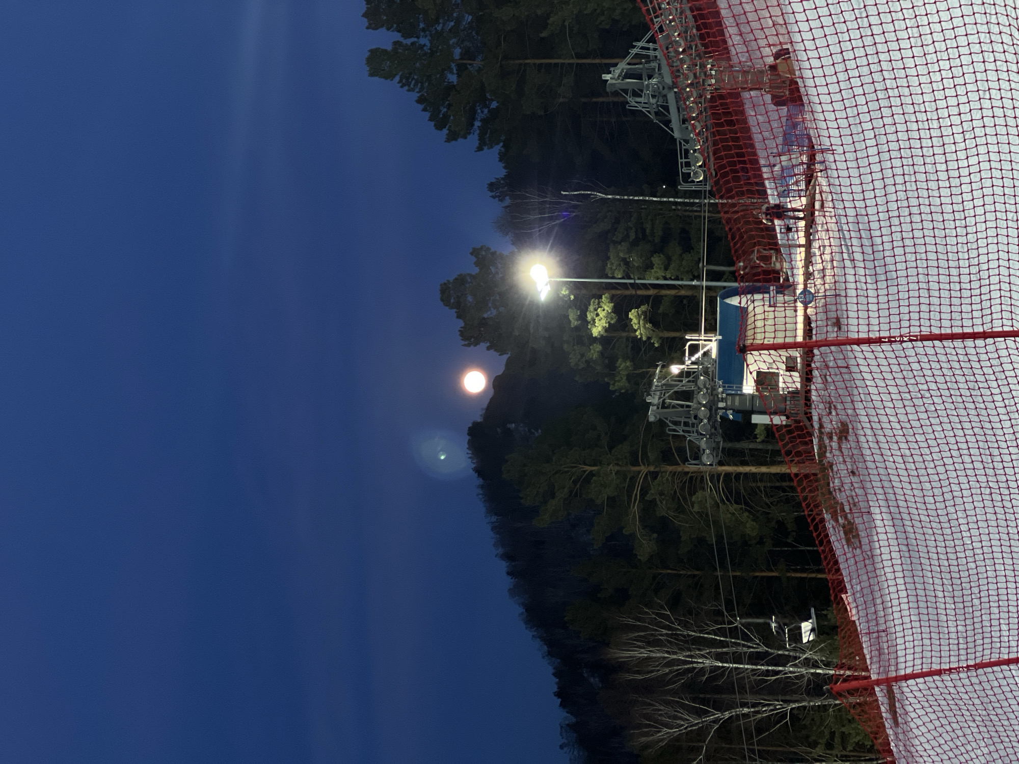 Here is a detailed description of the image:
The image shows a night scene at what appears to be a ski resort, potentially located in a forested area, illuminated by artificial light and possibly moonlight. The lower right portion of the frame is dominated by a red safety net covering a snow-covered slope. In the background, there are dense trees, which may be evergreen, extending upward into the frame. In the center of the image, a lamppost casts bright light onto the surroundings. The sky is a dark blue, and a bright round light is visible in the sky, probably the moon. Ski lift infrastructure is visible in the upper portion of the frame, with bare trees among evergreen. The orientation of the image suggests that the camera may have been tilted or the picture was taken sideways. There are no people visible in the image. The location is unknown.