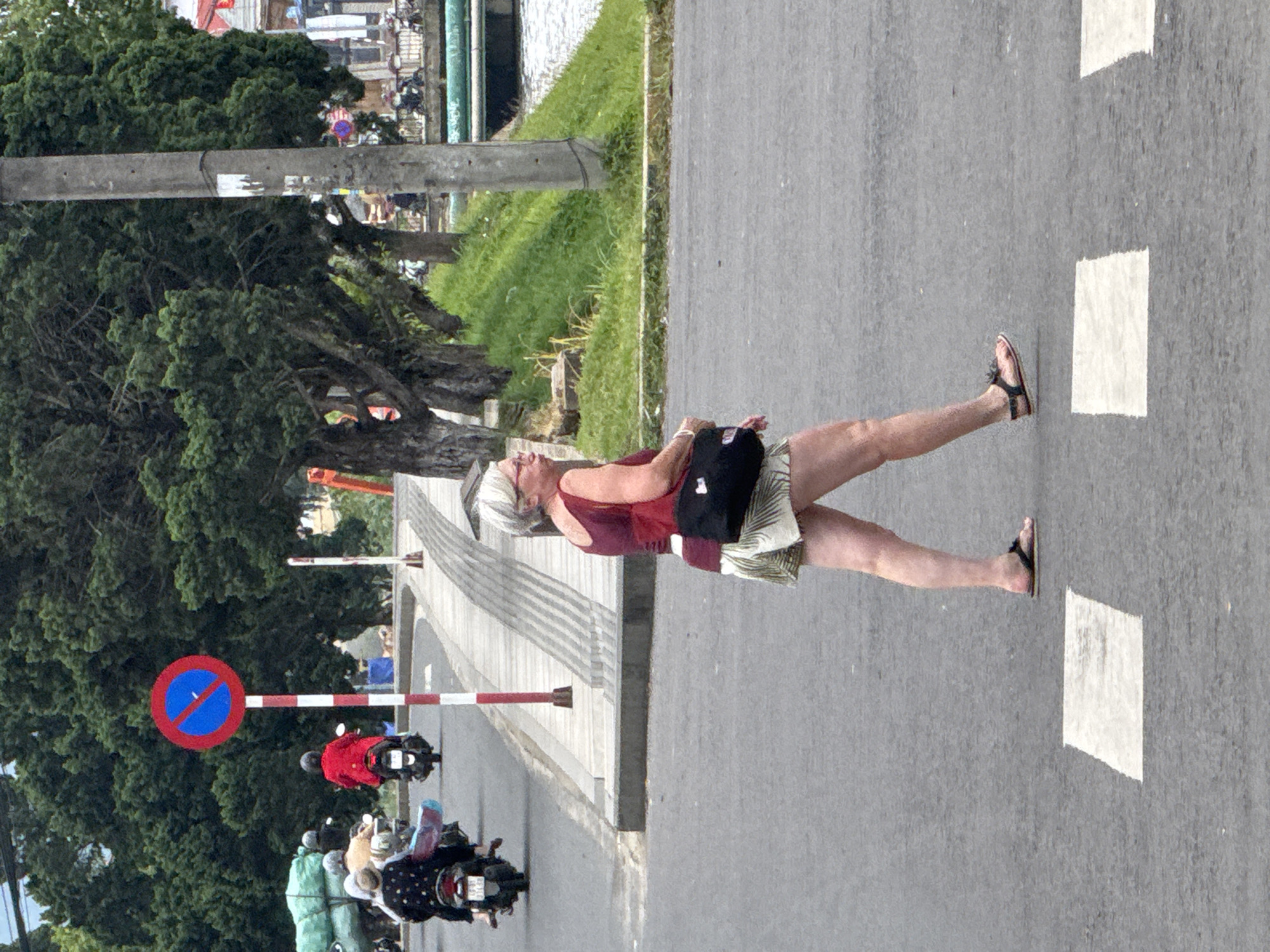 In the city of Phường Xuân Hương - Đà Lạt, Vietnam, a Caucasian woman with short grey hair and glasses is crossing a street at a pedestrian crossing. She is wearing a red sleeveless top, a light-colored patterned skirt, and dark sandals. A black fanny pack is slung across her right hip. She is walking from left to right in the frame. In the background, several motorbikes are visible, with one rider in a red jacket. A red and white striped pole with a circular "no parking" sign is positioned on the sidewalk to the left of the pedestrian. Lush green trees line the street, and a canal or body of water is visible on the right side of the frame, separated by a grassy embankment and a concrete structure. The street appears to be paved with asphalt.