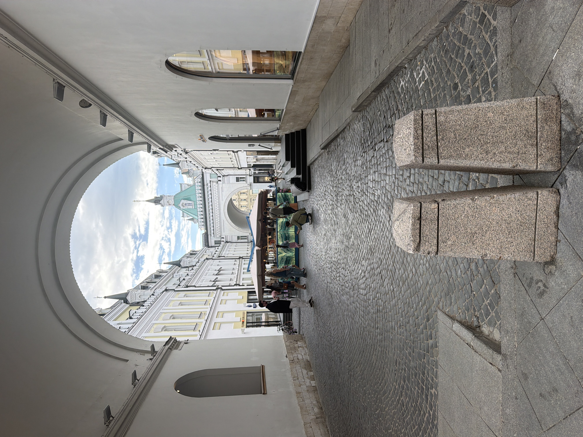 The image shows a view of a street in Moscow, Russia, framed by a large arched passageway. The architecture is predominantly white and yellow, with intricate details and spires visible in the background. People are present, either walking along the cobblestone street or seated at tables under an awning, which appears to be part of an outdoor café. In the foreground, there are two large, rectangular stone blocks on the right side of the image. The sky is visible through the arch, with scattered clouds. The street is paved with cobblestones. The building on the left side of the image has a series of arched windows and is painted white.