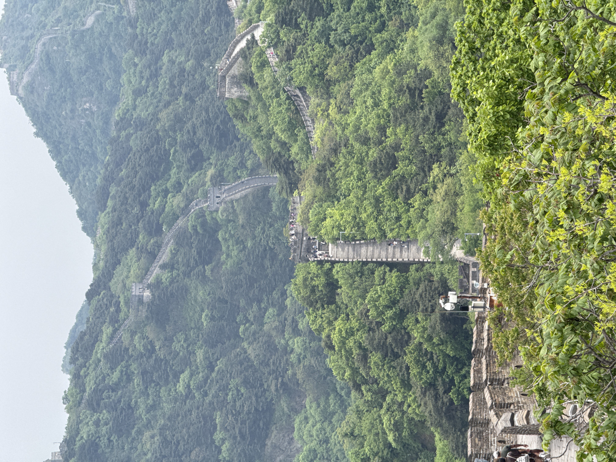 This aerial image depicts a section of the Great Wall of China in Huairou District, China. The wall winds through lush, green, mountainous terrain. Sections of the wall appear well-preserved, with visible brickwork and fortifications. Tourists are walking along the wall in some areas. The environment is predominantly green due to the dense foliage of the mountains. The sky is overcast. Trees surround the visible portion of the wall. There are CCTV cameras on the wall.