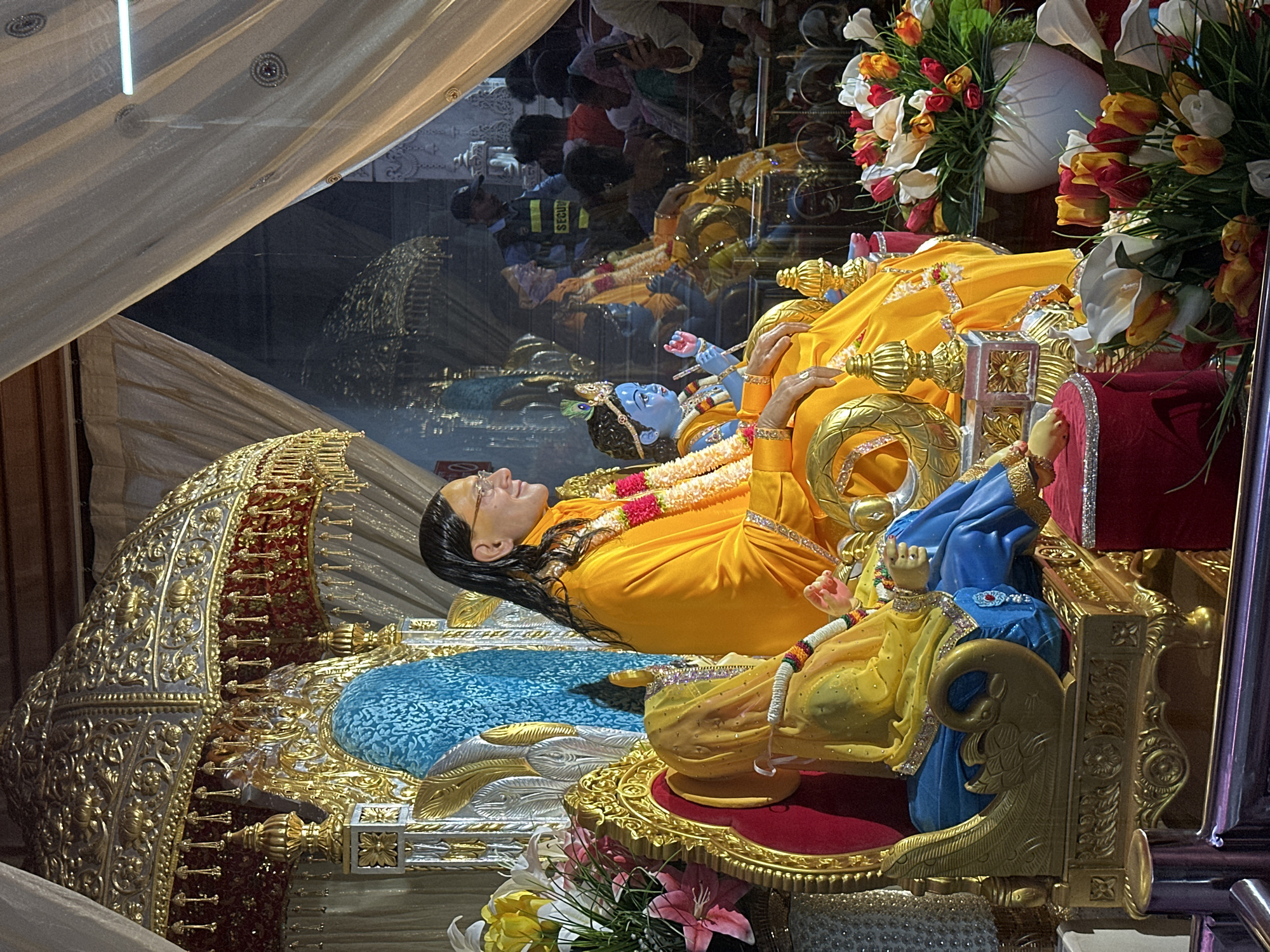 The image shows a deity of a person lying down, possibly in repose, and a smaller blue-skinned deity beside them. Both figures are adorned in bright yellow clothing and jewelry. The main figure has dark hair and glasses and appears to be resting on an elaborately decorated bed or platform with gold and blue accents. To the side, another figure in blue and yellow attire sits.
The scene is framed by a clear barrier, possibly glass, which reflects a crowd of people, including a security guard. This suggests the deities are in a public display or religious setting. The arrangement is further adorned with flowers, including lilies and tulips.
The image likely depicts a deity scene at the Prem Mandir temple in Chhatikara, India, known for its elaborate displays.
