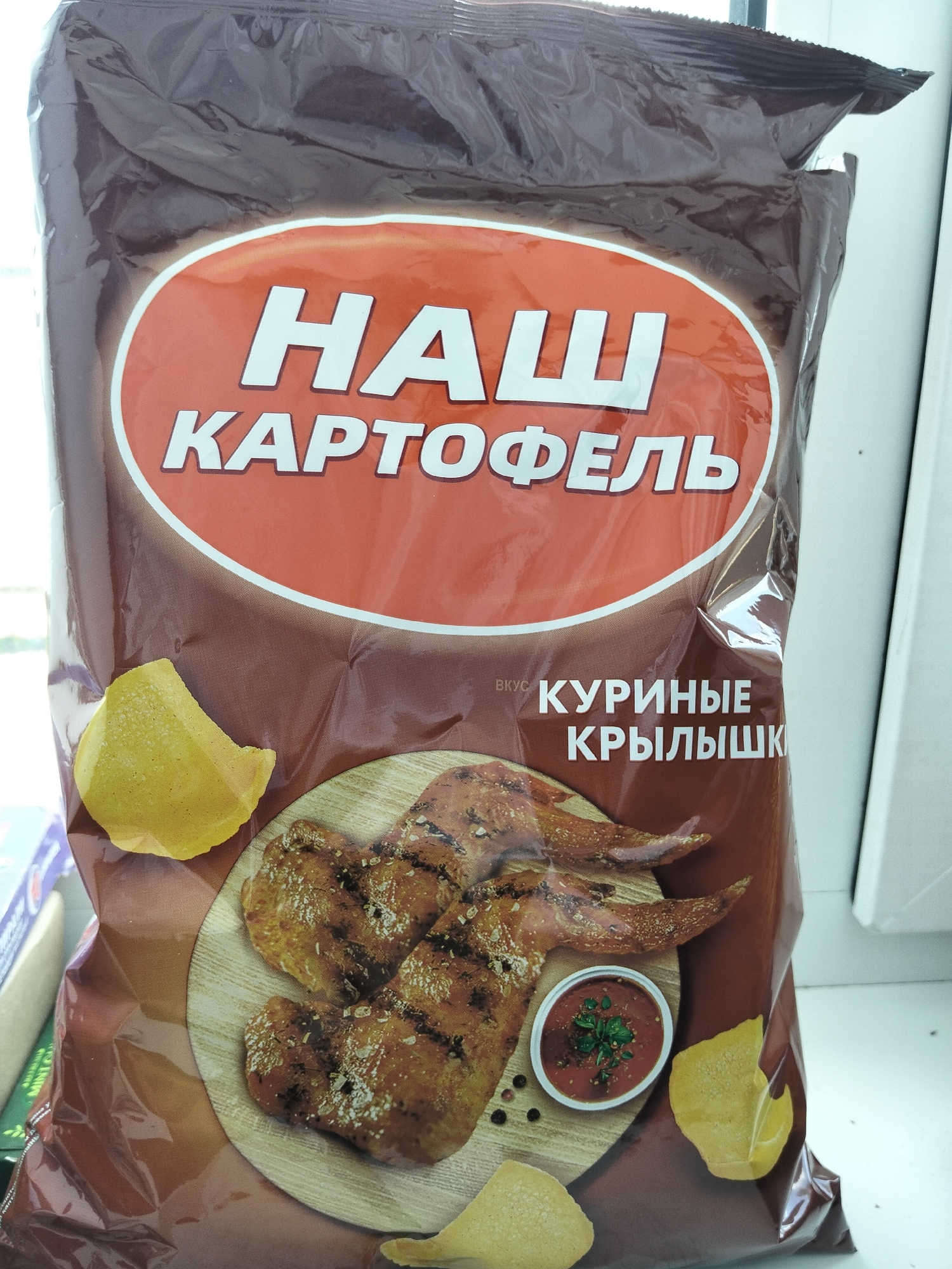 The image shows a bag of potato chips with the brand name "НАШ КАРТОФЕЛЬ" (Our Potato). The flavor is "КУРИНЫЕ КРЫЛЫШКИ" (Chicken Wings), as indicated by the text and the image of grilled chicken wings and a dipping sauce on the bag. The bag is dark brown and has a large oval emblem with the brand name in white lettering. The chips are depicted as golden yellow and crispy. The bag is positioned on a white surface, possibly a windowsill or counter, with a blurred background that suggests an indoor setting. There are no people or animals visible in the image. The lighting appears to be natural, coming from a window to the right of the bag. The location is specified as Oktyabrsky, Russia.