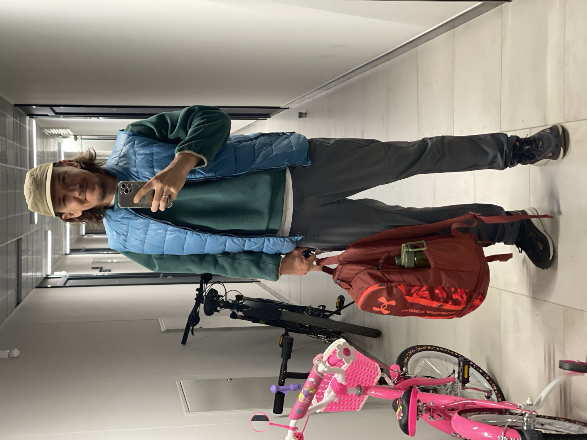 The image, rotated clockwise, shows a man taking a mirror selfie in an indoor hallway. The man has long brown hair and is wearing a beige baseball cap, a dark green fleece sweater over a white undershirt, a light blue puffer vest, dark grey pants, and dark athletic shoes. He holds a silver smartphone in his right hand, capturing his reflection. A wedding band is visible on his left ring finger.
He carries a red-orange backpack, which prominently features the "Under Armour" logo on its front, and a green water bottle is tucked into a side mesh pocket. The background reveals a clean, well-lit corridor with white walls and a light-colored tiled floor. Two bicycles are parked against the wall: a black adult bicycle and a smaller, bright pink child's bicycle equipped with training wheels, a pink basket, and tires adorned with heart patterns. The man has a relaxed expression with a slight smile. The setting appears to be a residential building hallway or common area, given the presence of both adult and child bicycles.
