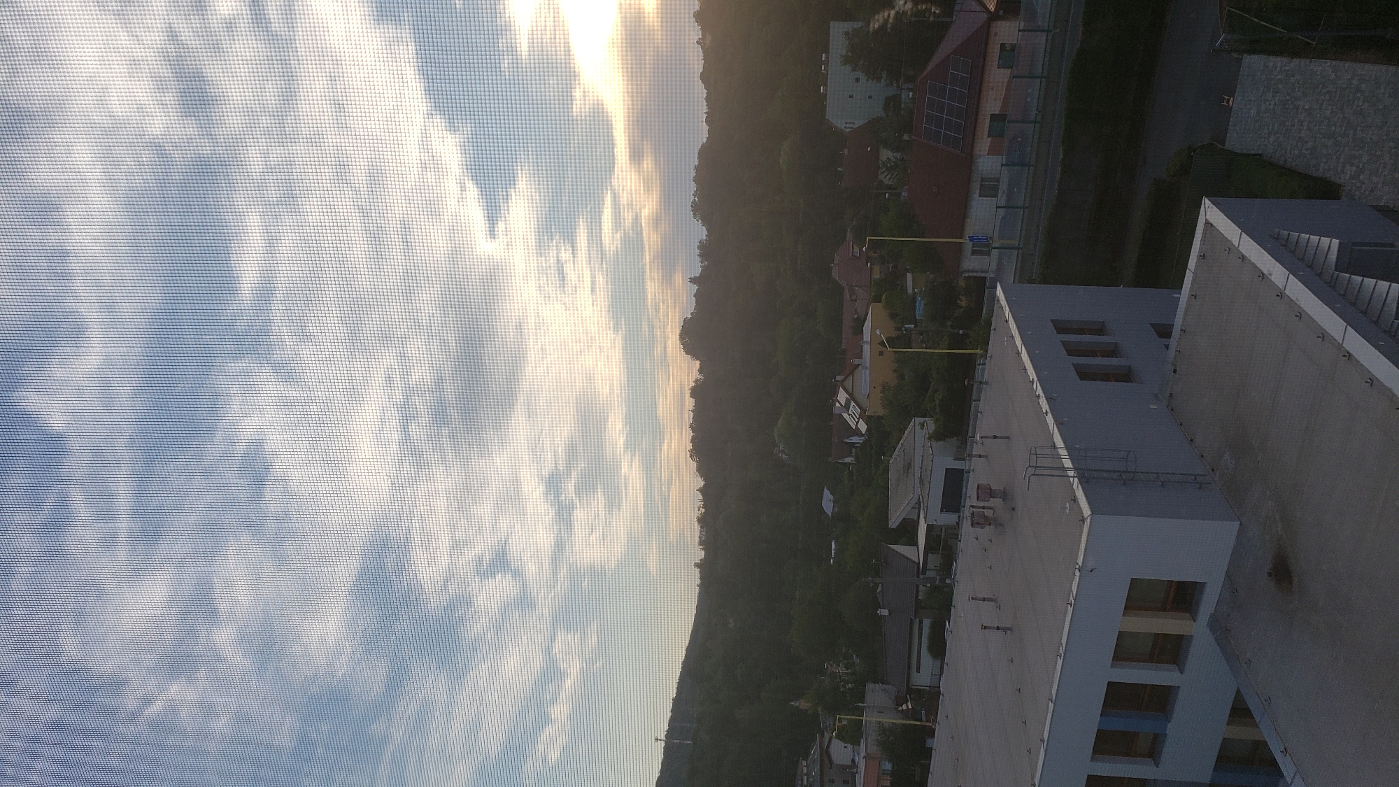 The image depicts a view from a window with a screen overlay, looking out over a residential area in Prešov, Slovakia. The sky is partially cloudy with the sun setting or rising on the right side, casting a warm glow. In the foreground, the roof of a modern, multi-story building with a light grey facade and rectangular windows is visible. The building appears to be a school or office complex, with ventilation pipes and a ladder on its roof. Beyond this building, a suburban landscape unfolds with numerous houses, most of which have pitched roofs, some with solar panels. A road runs through the middle ground, with streetlights and a fence separating it from the houses. A densely wooded hill forms the backdrop, suggesting the city is nestled within a natural landscape. The overall impression is one of a quiet, everyday scene in a European city.