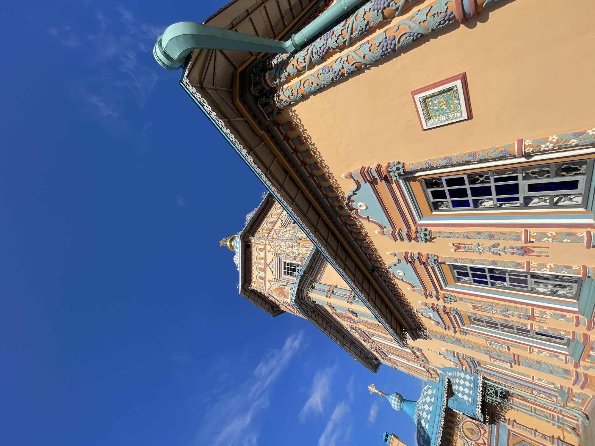 An upward-angled view captures the exterior of a vibrantly decorated building, likely a church or cathedral, against a clear azure sky. The structure is characterized by its ornate and colorful architectural style, featuring walls painted in a warm yellow or ochre hue. Numerous decorative elements adorn the facade, including intricate bas-reliefs of grapevines with purple grapes and green leaves, floral patterns, and geometric designs in shades of blue, light green, maroon, and white.
A distinctive light blue/teal gutter system runs along the eaves, which are also embellished with white, lacelike carvings. Multiple windows are visible, some with clear panes and others appearing to have dark blue or reflective glass, all framed by colorful decorative borders. A small, square, embedded tile-like artwork depicting yellow birds or motifs on a light green background with a maroon frame is also present on the wall.
In the background and lower right, additional elements of the building are visible, including a golden onion dome and a blue-and-white checkered cupola, both topped with golden Orthodox crosses. The sunlight is bright and direct, casting clear shadows that emphasize the three-dimensional texture of the decorations. The sky is largely cloudless, save for a few wispy white clouds. No people or activities are depicted, and no discernible text is visible on the building. The architectural style suggests Eastern European or Russian Orthodox design.