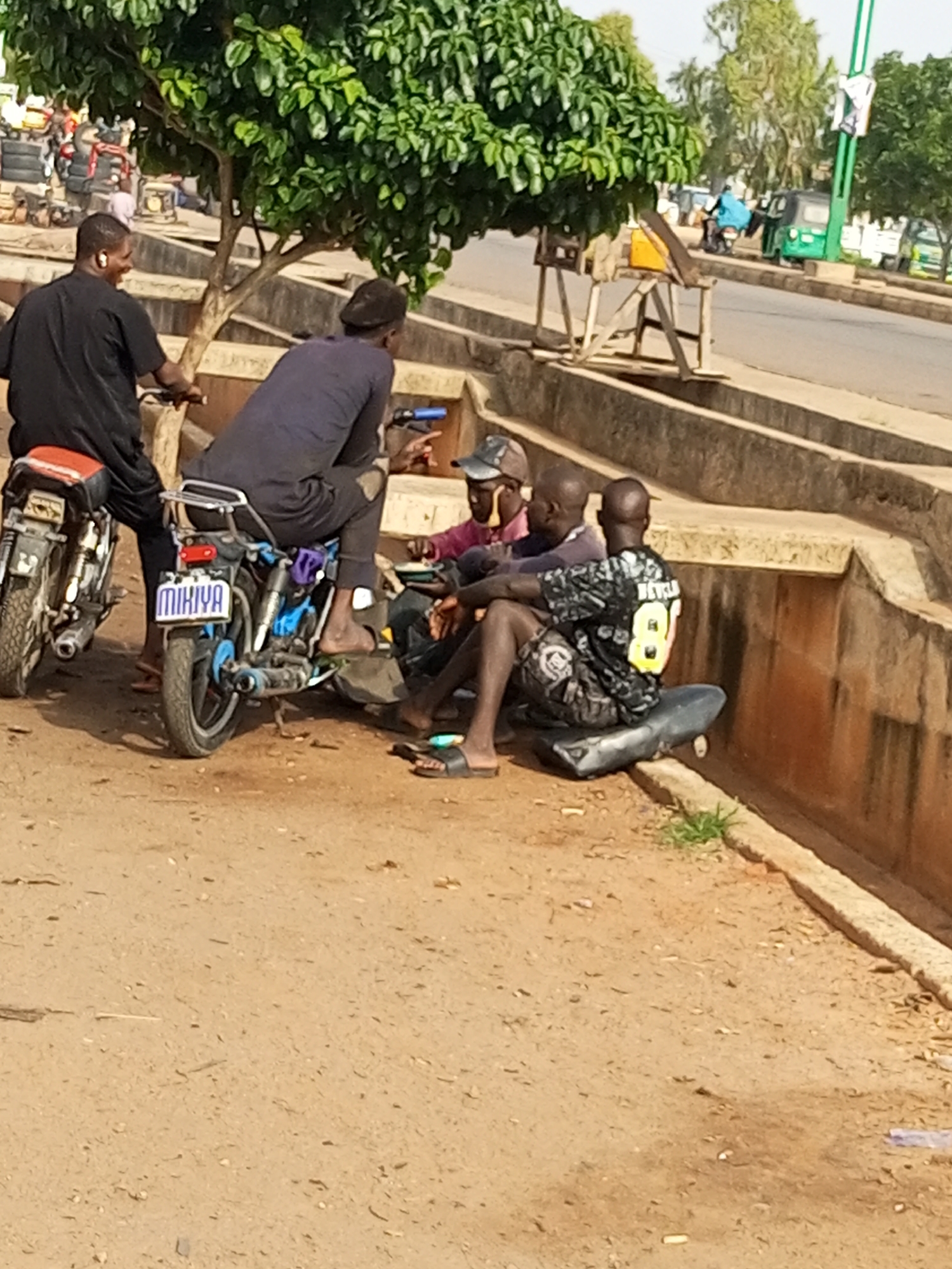 In Wuro Dole, Nigeria, four individuals are observed near a dirt road adjacent to a concrete embankment and a paved road. In the foreground, a person wearing a black t-shirt and dark pants is seated on a motorcycle. Another person, dressed in a black t-shirt and dark pants, is also seated on the same motorcycle, facing away from the camera. To their right, a third individual in a dark blue shirt and dark pants is seated on another motorcycle. The fourth person, wearing a pink shirt and a cap, is seated between the two motorcycles. The motorcycles are positioned close to each other. The environment appears to be an urban setting with a tree and some vegetation present.