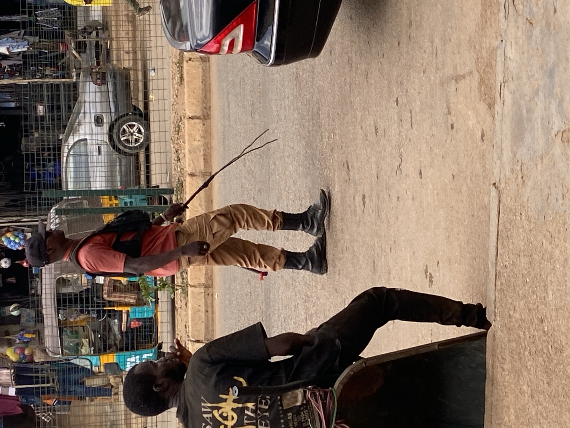 The image shows a street scene in Bukuru, Nigeria. A man wearing a red shirt, khaki pants, black boots, and a black backpack is walking with a thin branch in his hand. He is wearing a black hat. In the foreground, another man wearing a black shirt is partially visible, leaning into a container. A car is partially visible in the top center of the image. The background shows a metal structure filled with various objects. The street is paved, and a wall is on the right side of the image.