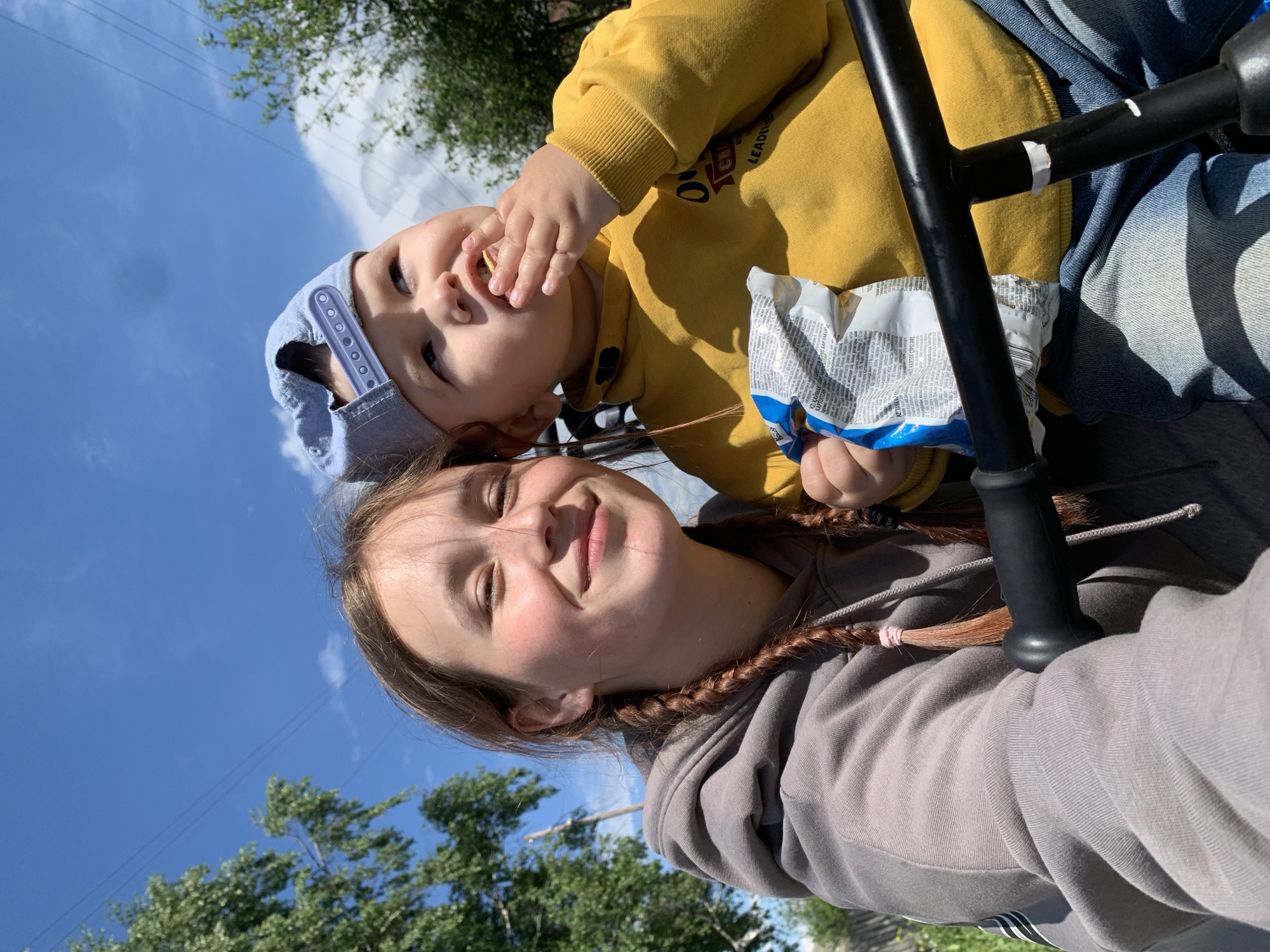 Two individuals are visible in an outdoor setting. The foreground is dominated by an adolescent female, wearing a grey hooded sweatshirt and sporting braids tied with pink bands. Behind her, a toddler wearing a yellow sweatshirt and a light blue cap is eating from a bag of chips. The setting seems to be outdoors, with trees and a blue sky with some clouds visible in the background. Based on visual cues, there's no specific indication that would confirm the location as Фоки, Russia.