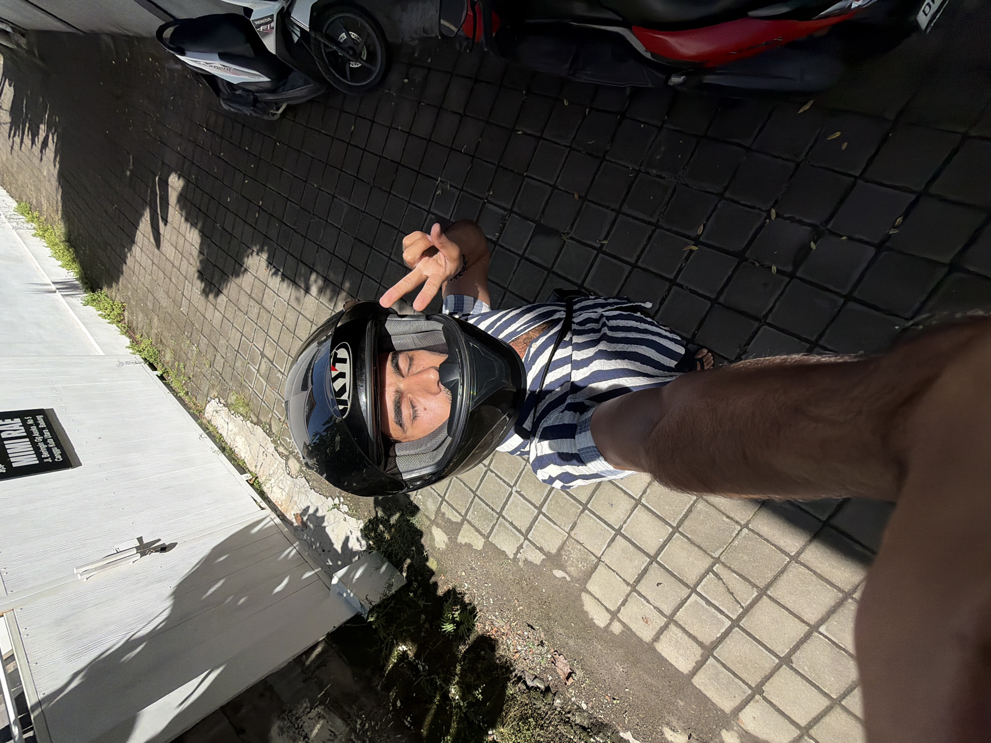 The image is a high-angle selfie taken by an individual wearing a black KYT motorcycle helmet and a striped blue and white shirt. The person is flashing a peace sign with their right hand, which is positioned near the helmet.
The selfie is taken on a paved area constructed with concrete paving stones. Two motorcycles are visible in the background, one primarily white and the other red and black. The presence of vegetation suggests the location is outdoors.
A partially visible sign indicates the location is "MIMI RAE, Jl. Beringin Gg Asoka. No 5, Canggu Kuta Utara, Badung." Thus, it is set in North Kuta, Indonesia.