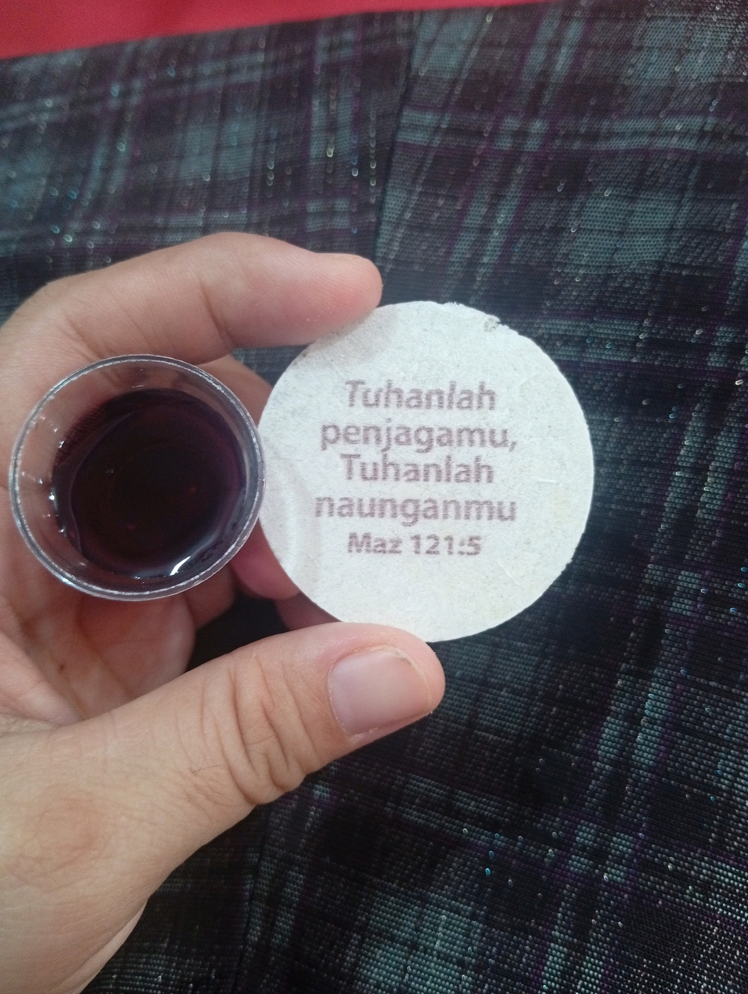 A close-up shot shows a hand holding a small, clear plastic cup filled with a dark reddish-purple liquid. Next to the cup is a round, white paper lid with text printed on it. The text appears to be in Indonesian and reads: "Tuhanlah penjagamu, Tuhanlah naunganmu Maz 121:5". This translates to "God is your keeper, God is your shade Psalm 121:5". The background is a dark, patterned fabric with a subtle sheen. The image is a candid shot, suggesting it was taken in a casual setting. No specific location in the city of Gunungsitoli, Indonesia can be determined from the image.