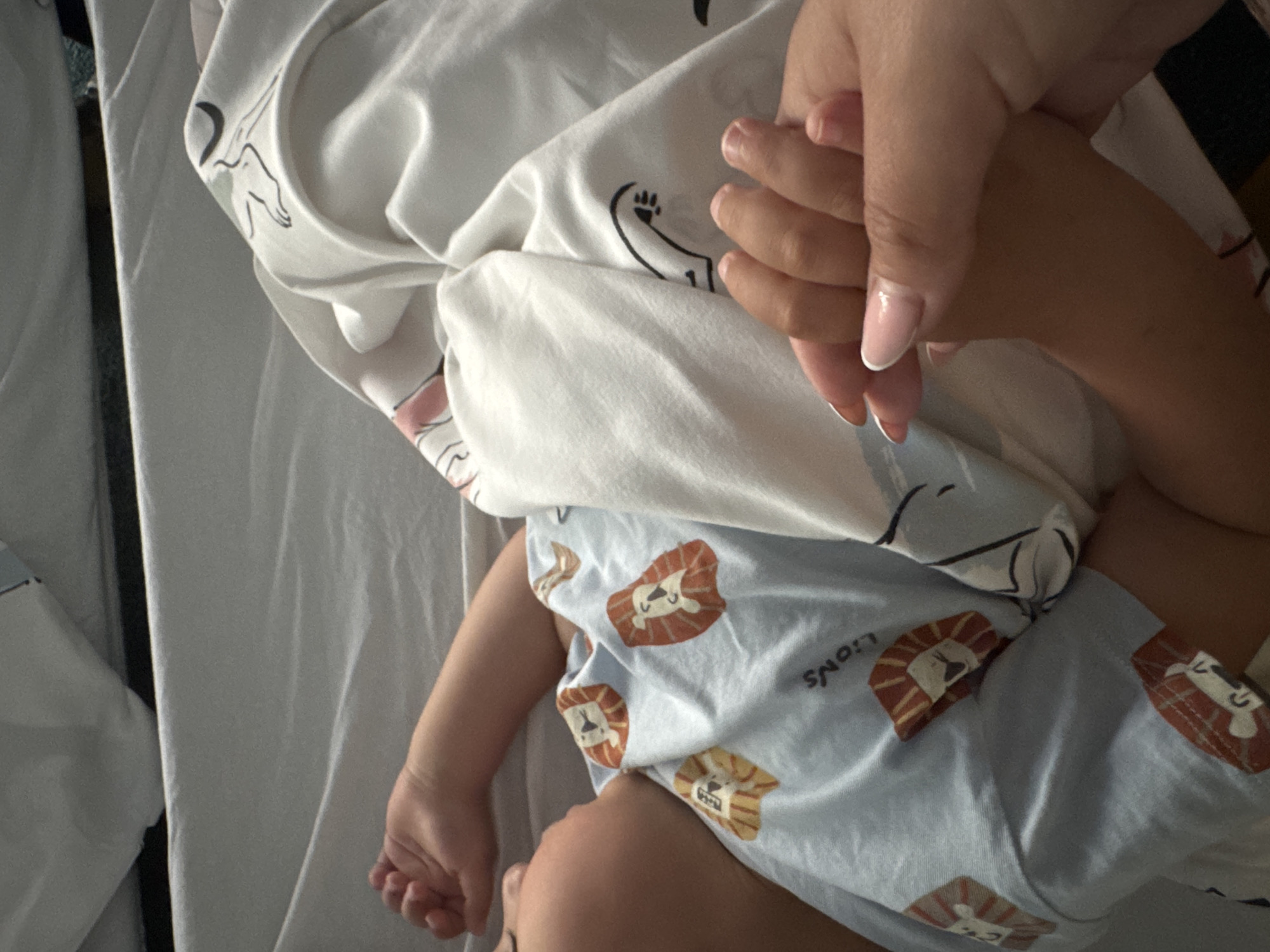 The image displays an intimate moment between an adult and a baby or young child, viewed from an overhead perspective. An adult's hand, with well-maintained nails featuring a pink base and white French tips, is gently holding the small hand of the child.
The child is depicted lying down, with a visible arm, leg, and a portion of their head. They are dressed in a light blue garment adorned with multiple cartoon lion faces, some of which clearly display the word "LIONS" in black text.
The setting appears to be a bed or cot, covered with white bedding. A plain white sheet is visible, along with a white blanket or duvet that features faint black line drawings, including a distinct paw print and other abstract, stylized shapes. The soft, even lighting suggests an indoor environment, possibly a bedroom, creating a warm and tender atmosphere.
The interaction between the adult and child, through the gentle clasping of hands, conveys feelings of love, comfort, and security. The scene is static, capturing a quiet moment of connection. The location context suggests this scene is taking place in Sofia, Bulgaria.