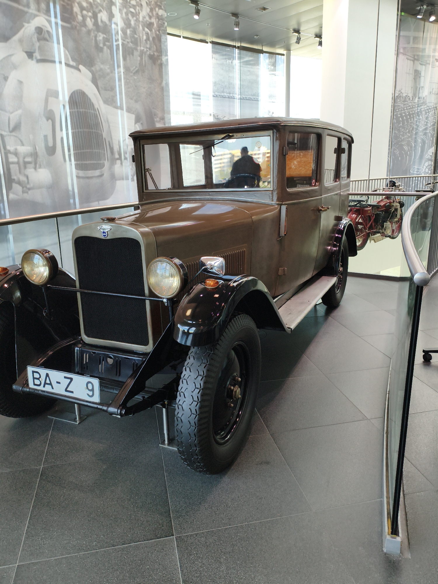 The image shows a brown, vintage four-door sedan parked indoors, likely in a museum. The car has a prominent grille, two large headlights, and a license plate that reads "BA-Z 9". A person is visible inside the car, seated behind the steering wheel. In the background, there is a large black and white mural depicting a racing car and a crowd. To the right of the car, a red motorcycle is displayed. The flooring is dark gray tile. The overall setting suggests an exhibition or display area within a public building. While the specific location is not evident in the image, the presence of a vintage car and historical imagery might suggest a museum dedicated to automotive history. Given the context of the question referring to Ingolstadt, Germany, this could potentially be the Audi museum mobile, which features historic Audi vehicles.
