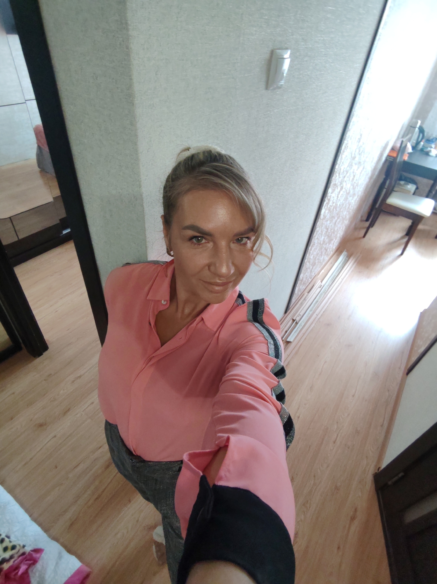 A medium shot shows a blonde woman with her hair pulled back in a bun, taking a selfie in what appears to be an indoor hallway. She is wearing a pink button-down shirt with metallic stripes on the sleeves and dark pants. The hallway has light-colored textured wallpaper on one side, and a wooden floor. To the right, a dark-colored doorway is visible, and further down the hall, a table and chair are partially visible. The woman's left shoulder and upper arm are visible as she holds her phone to take the photo. There is no identifiable information to place this image in Petropavlovsk-Kamchatsky, Russia.