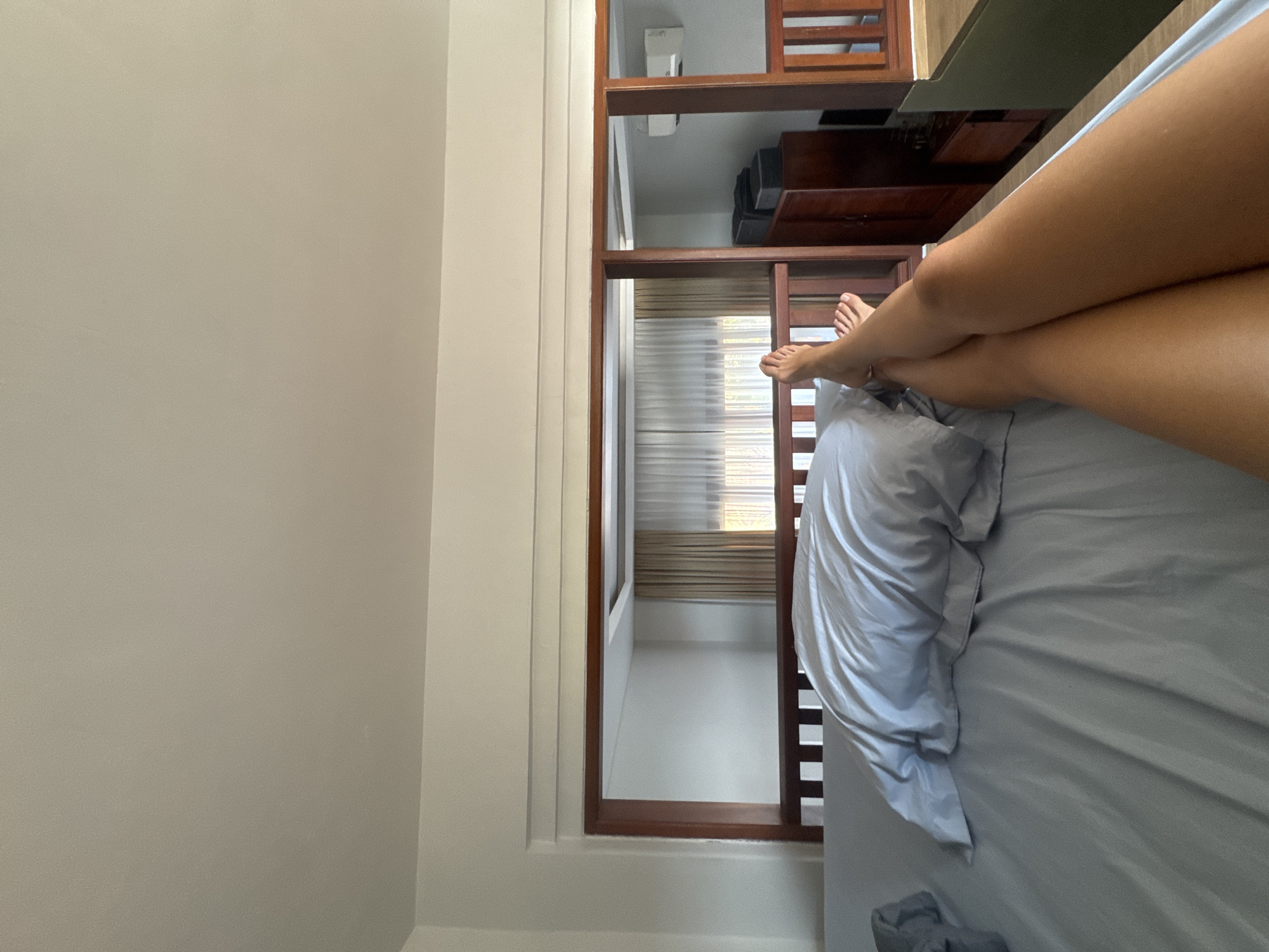A first-person perspective shot shows a person's legs and feet resting on a bed in what appears to be a hotel room in North Kuta, Indonesia. The bed is covered in light blue sheets and a rumpled duvet. The headboard of the bed is made of dark wood with vertical slats. Beyond the bed, there is a wooden frame that creates an opening, revealing another section of the room. This area includes a closet with dark wood doors, a television, and an air conditioning unit mounted on the wall. Sheer curtains hang in front of a window, allowing soft light to filter into the space.