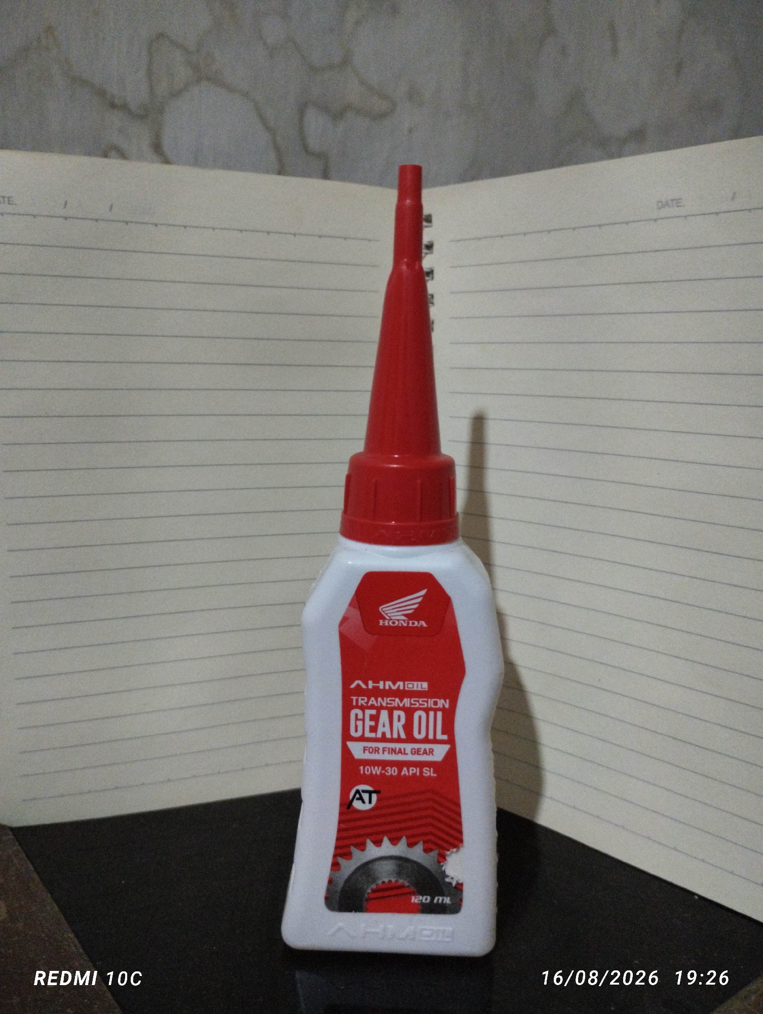 A bottle of Honda Gear Oil for final gear with 10W-30 API SL specification sits on a table next to a notebook. The bottle is white with a red cap and label, featuring the Honda logo and the words "TRANSMISSION GEAR OIL." The notebook has lined pages with the word "DATE" printed at the top. The background is a textured wall. A timestamp in the bottom right corner indicates the image was taken on August 16, 2026, at 19:26. The text "REDMI 10C" is present in the bottom left corner, indicating the device used to capture the image. There are no people or interactions depicted. This image is a product photograph, not indicative of any specific location within Kembiritan, Indonesia.