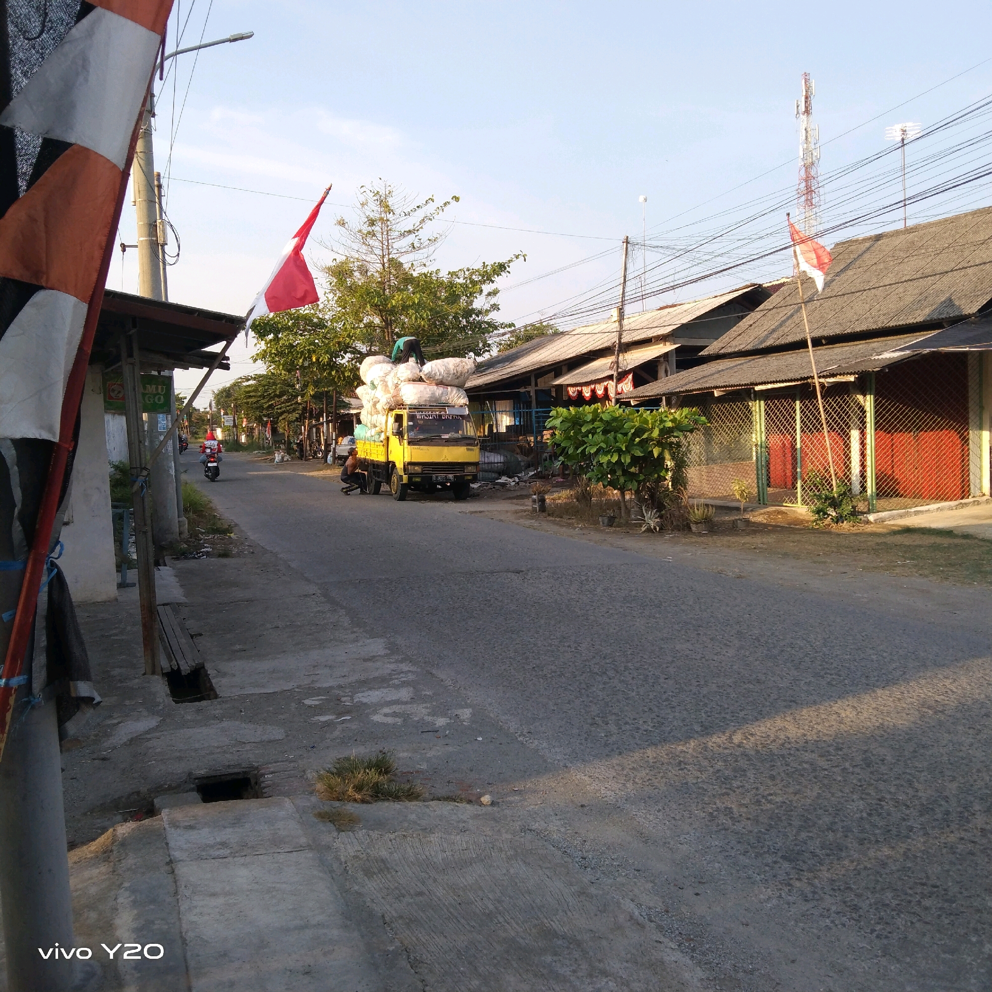 In Rawagempol Wetan, Indonesia, an outdoor scene unfolds on a paved road lined with buildings and vegetation. A yellow truck, heavily laden with large white sacks, is parked on the right side of the road. Two individuals are visible near the truck: one is sitting on the ground near the front of the truck, and another is standing on top of the sacks, appearing to adjust them. A red and white Indonesian flag flutters from a pole near the truck. Further down the road, a person rides a motorcycle. On the left side of the road, a street lamp and power lines are present, along with a building that has a covered entrance. Flags are also visible on the left side of the road, attached to a pole. The sky is clear and a soft light suggests it might be morning or late afternoon.