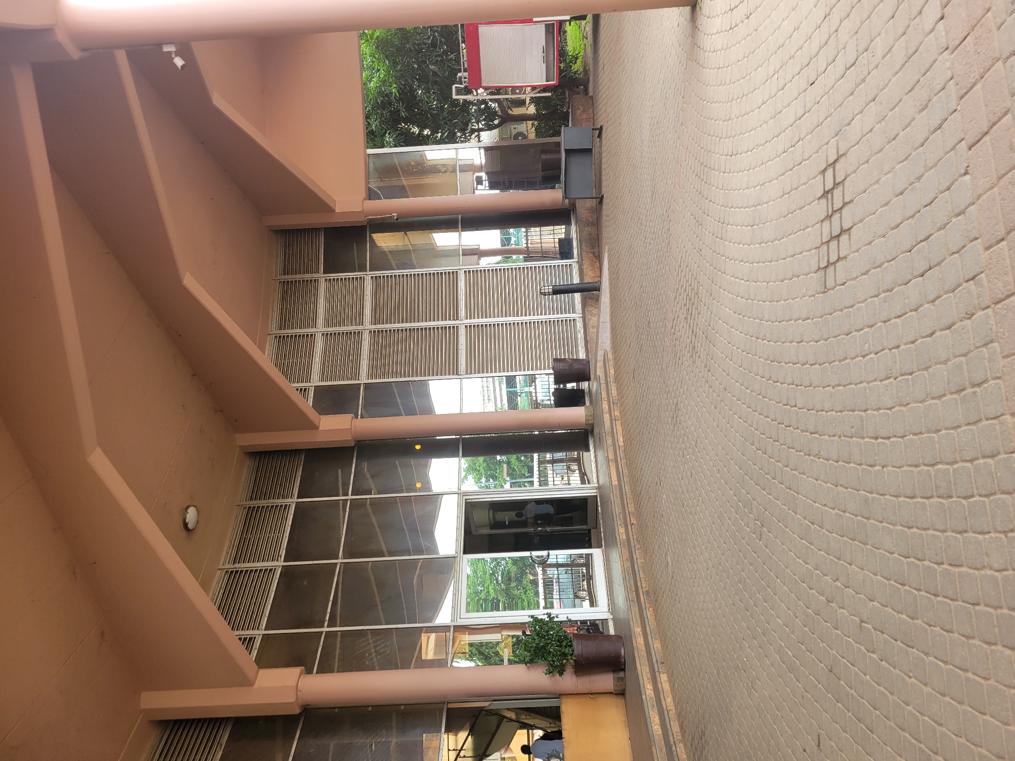 This image, set in Abuja, Nigeria, captures the exterior of a modern building and an adjacent paved walkway during bright daylight. The building features a light peach or salmon-colored facade with pronounced structural beams creating an overhang. The lower part of the building is dominated by large glass panels, some of which have horizontal slatted sections, possibly for ventilation or aesthetics. A main entrance with glass doors is visible, reflecting the surrounding greenery and sky.
The expansive walkway is paved with light-colored, possibly beige or off-white, interlocking blocks arranged in a distinct, sweeping radial or curved pattern. A dark, rectangular wet or stained patch is noticeable on these pavers.
Several objects are present: two large, dark potted plants flank the main entrance, adding a touch of greenery. A dark grey outdoor waste bin or receptacle stands on the right side of the walkway near the building. Mounted on the building's facade and under the overhangs are several small white light fixtures or security cameras. In the upper right background, partially obscured by trees, is a red rectangular structure with a white panel, possibly a utility box or signage, and a white outdoor air conditioning unit.
Reflections in the glass doors suggest activity, with a person in a light-colored shirt visible through the entrance, indicating people potentially entering or exiting the building. The overall scene suggests a commercial or office building, or possibly a hotel entrance. The time of day appears to be daytime with clear weather, judging by the bright illumination and visible shadows. No discernible text is clearly readable in the image.