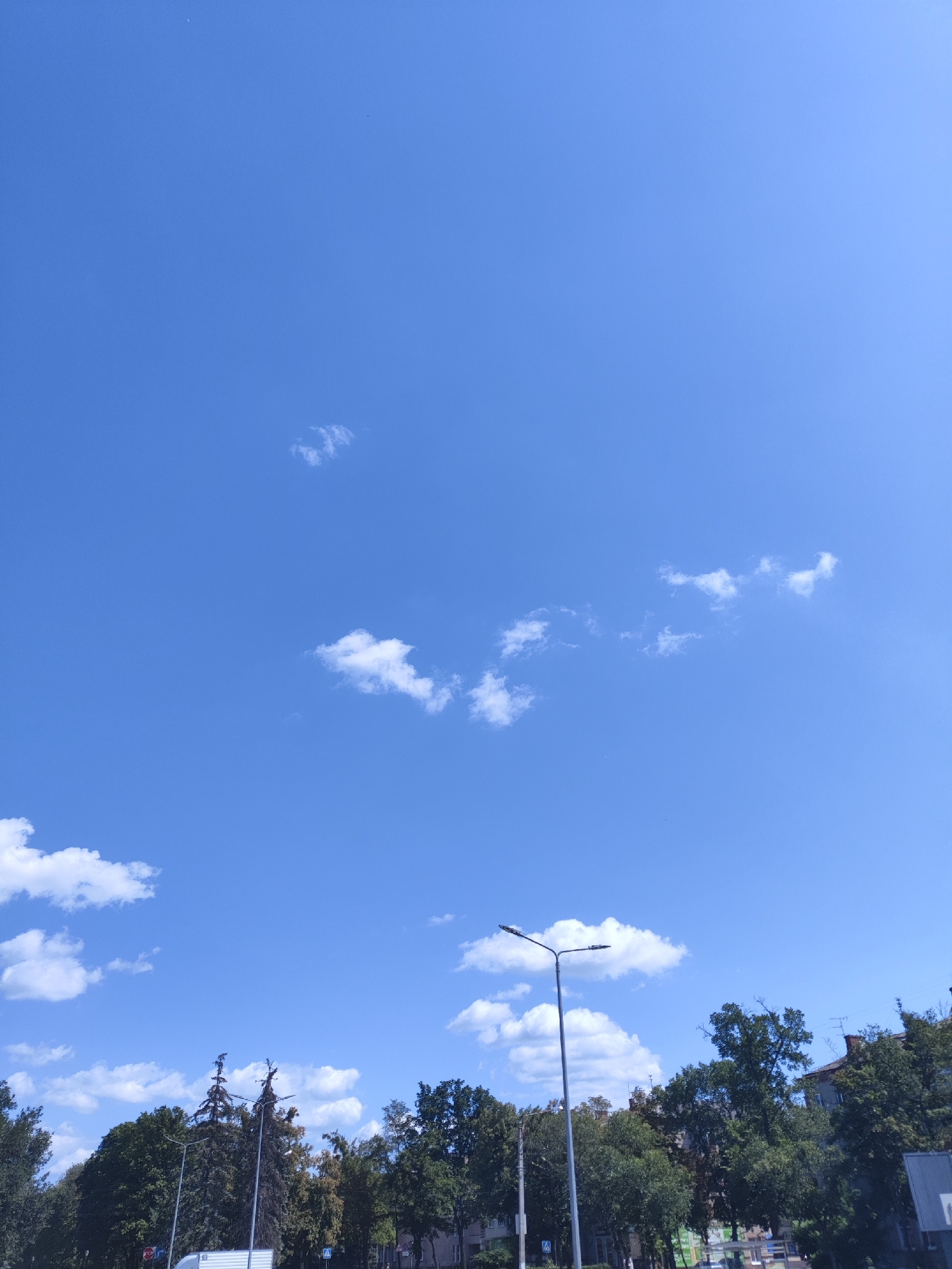 The image captures a clear blue sky with scattered white clouds over a city landscape. In the foreground, a line of trees is visible, with taller trees in the center and to the left, and buildings and shorter trees to the right. A street light stands prominently on the right side of the image, and a stop sign is visible on the left. The overall scene appears to be a daytime view of an urban environment, possibly a street or park area in Kharkiv, Ukraine, characterized by greenery and a bright sky.