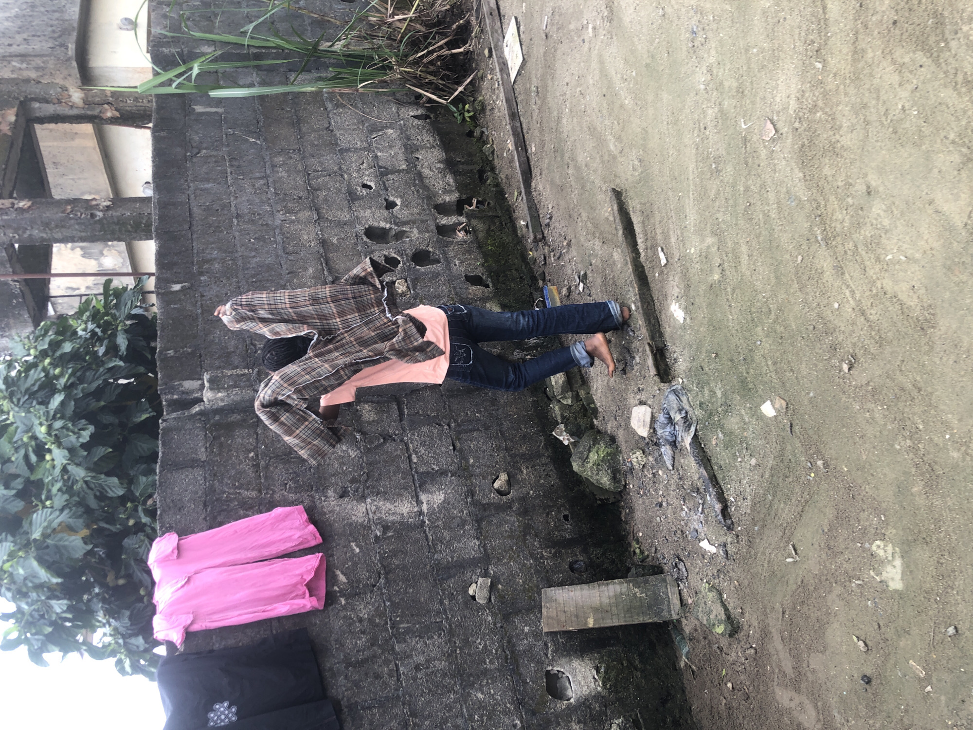 In Ado, Nigeria, a person is seen from the back, standing in front of a rough, grey, cinder block wall. They are wearing blue jeans and a light pink t-shirt. The person is in the process of taking off a plaid shirt, which they are holding up with both hands. To the left of the person, a black t-shirt and two bright pink shirts are hanging on a line against the wall. The ground in front of the wall is a mix of dirt and sand, with some debris scattered around, including rocks, pieces of wood, and plastic. Green foliage can be seen growing over the wall in the upper left portion of the image, and tall green grass is visible on the right side. The overall environment appears to be an outdoor, utilitarian space.