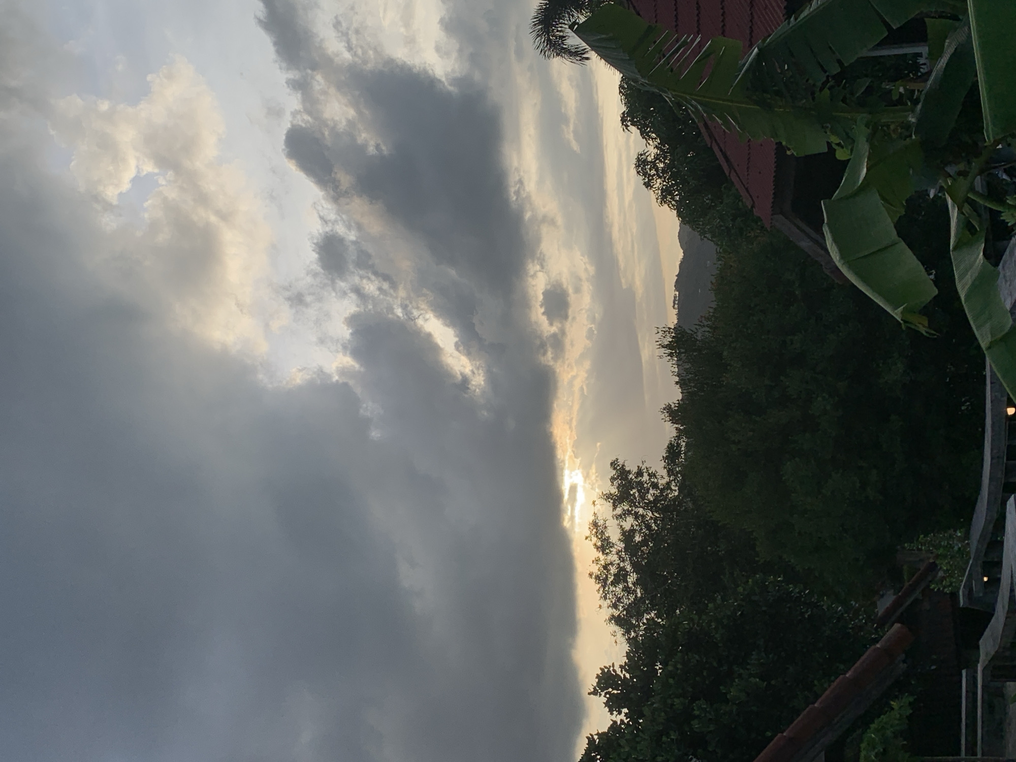 The image depicts a cloudy sky at dusk in บ้านบางรักษ์, Thailand. Heavy gray clouds dominate the left side of the frame, while the setting sun peeks through a gap between the clouds on the right, casting a golden glow. Lush green foliage, including trees and banana plants, occupies the right side of the scene, and a glimpse of rooftops and structures suggests a residential area. A mountain range is faintly visible in the distance.