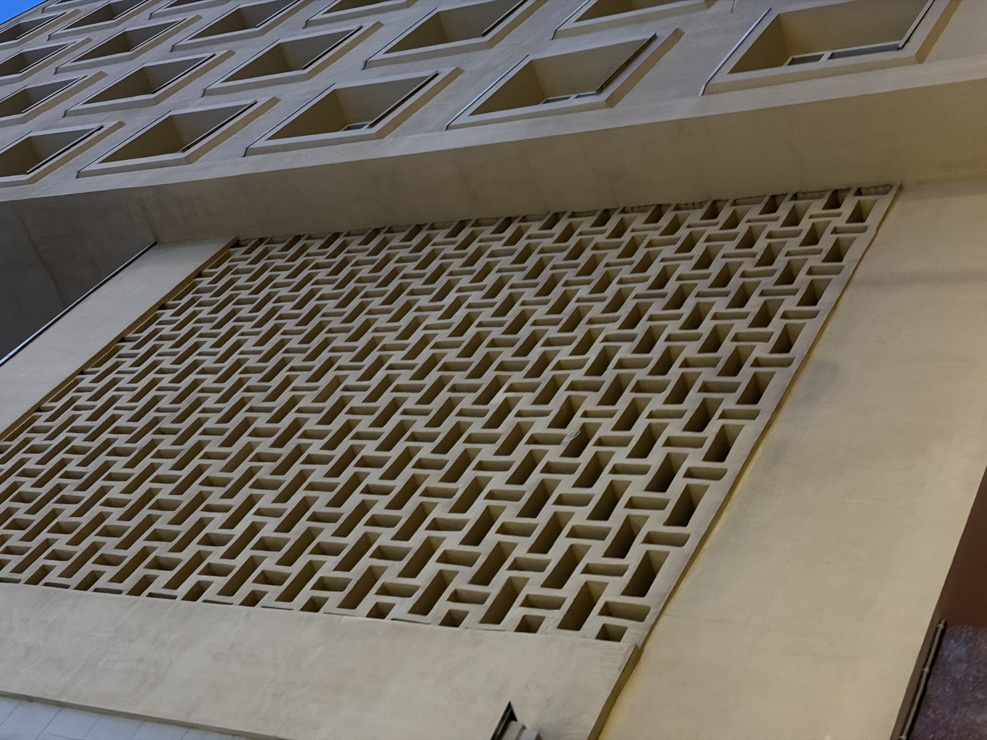 The image shows a low-angle view of a building facade, likely taken in Moscow, Russia. The upper portion of the image displays a section of the building's multiple windows and an overhang. Below this, a decorative, rectangular latticework panel is visible. The latticework is made of horizontal and vertical rectangular openings and creates a patterned effect. The colors are muted and predominantly beige or tan, and the lighting is subdued, with some shadows indicating a time of day when the light is not direct, possibly late afternoon or early evening. The overall composition is architectural, focusing on the building's design elements.