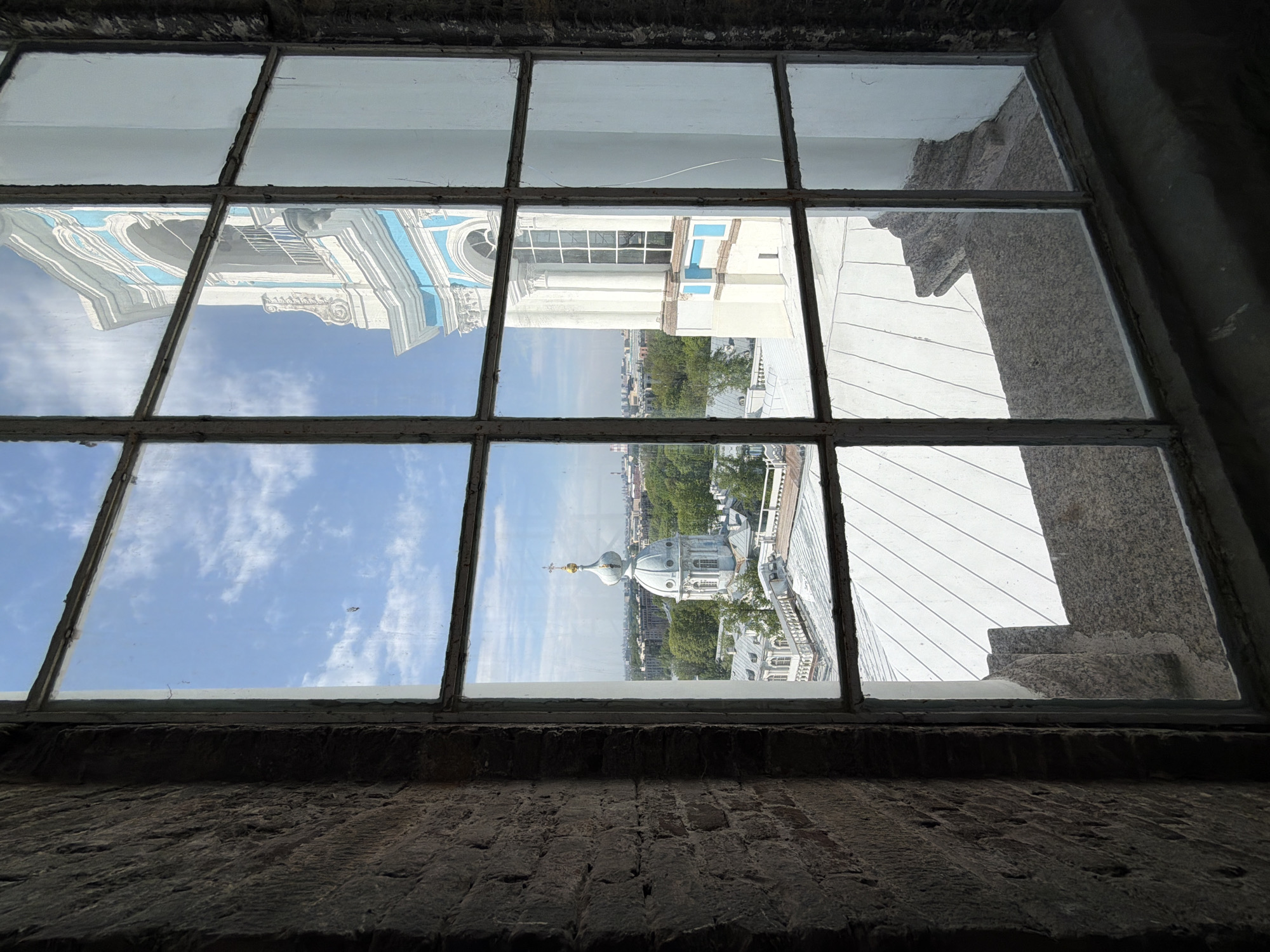The image depicts a view of Saint Petersburg, Russia, seen through a multi-paned window. The window frame is dark and appears aged, constructed of dark brick or stone. The reflections in the glass show buildings, including a church dome. The sky is visible through the glass, indicating a clear day. The buildings visible outside are upside-down due to the reflection in the window panes.