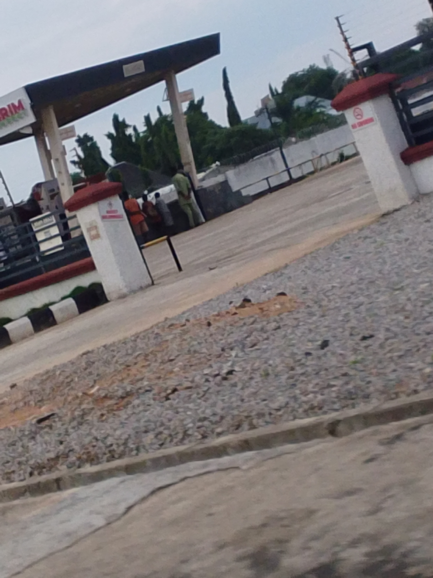 The image shows a gas station in Jalingo, Nigeria. The gas station has a distinctive angled roof supported by pillars. In the foreground, there is a gravel area and a paved road. A white pillar with a red top and a "no smoking" sign is visible. Behind the pillar, several people are gathered, some under an umbrella. One person appears to be wearing a light green uniform. The background includes trees, buildings, and a fence. The sky is overcast.