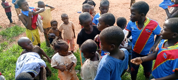 A group of approximately twenty children, estimated to range from toddlers to pre-teenagers (2-12 years old), are gathered outdoors in the city of Ngalula, Democratic Republic of the Congo. The ground features prominent areas of red-brown earth interspersed with patches of green grass, particularly in the lower left and central foreground. The children are dressed in varied attire; several wear light-colored t-shirts or dresses, some exhibiting dirt stains. Two children prominently display blue and red shirts with a diagonal red stripe and an emblem resembling the flag of the Democratic Republic of Congo. One boy in the upper left, wearing yellow shorts, holds blue and red/white fabric items. Another child in the lower left crouches or sits in the grass. The children are clustered closely together, with many observing each other or an unseen focal point just outside the frame, suggesting a collective gathering or interaction. The scene is illuminated by natural daylight.