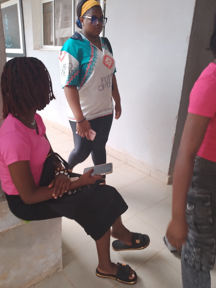 Three women are in an indoor corridor. One woman with dreadlocks is sitting on a concrete bench wearing a pink shirt and black skirt, holding a phone. Another woman is standing behind her, wearing glasses, a yellow headband, a white and turquoise patterned top, and black leggings, also holding a phone. The third woman, wearing a pink shirt and patterned jeans, is partially visible on the right side of the frame. The location is likely a building corridor in Ogboko, Nigeria.
