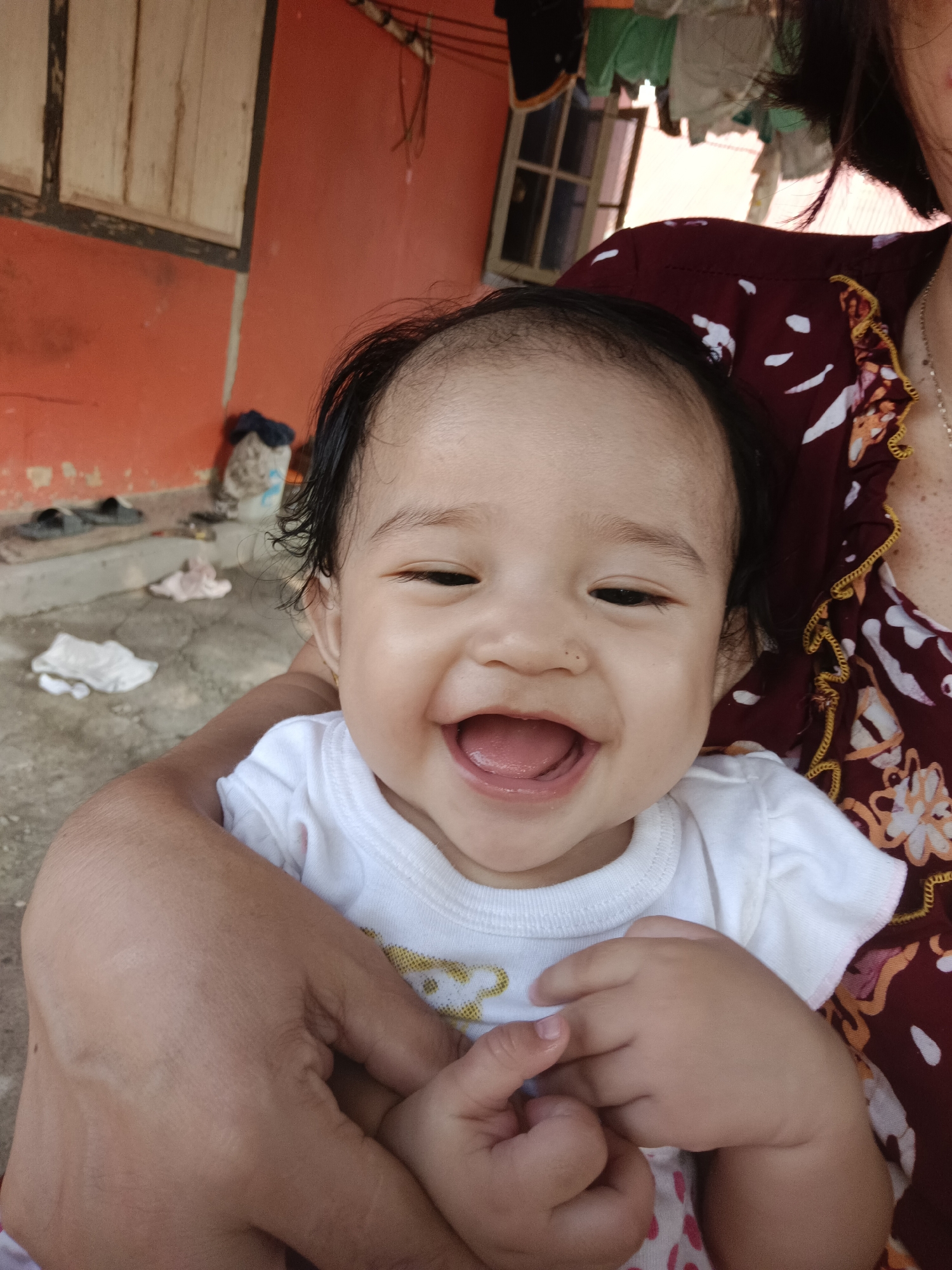 A close-up, waist-up shot shows a baby with dark hair, light skin, and dark eyes, laughing with an open mouth, showing its tongue. The baby is wearing a white onesie with a yellow graphic. The baby is being held by an adult whose arms are visible supporting the baby. The adult is wearing a dark red patterned garment with a yellow trim. The background appears to be outdoors, with an orange-painted wall, a wooden window frame, some shoes, and laundry hanging in the distance. The ground is concrete and appears to be a courtyard or outdoor living space. The setting is likely a residential area in Kawua, Indonesia, given the architectural style and outdoor elements.