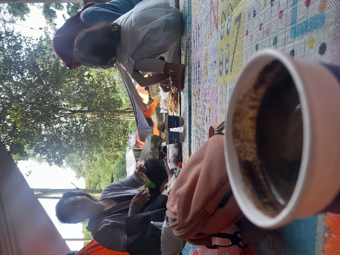 A campsite scene features a woman in a gray shirt and black top eating inside a tent. Near her is a brown backpack. A child with dark hair wearing a white shirt and purple hoodie sits on a colorful mat beside a woman. In the foreground, an out-of-focus mug of coffee sits on the mat. The background shows lush green trees. The photo appears to be taken from within the tent, capturing a casual outdoor moment.