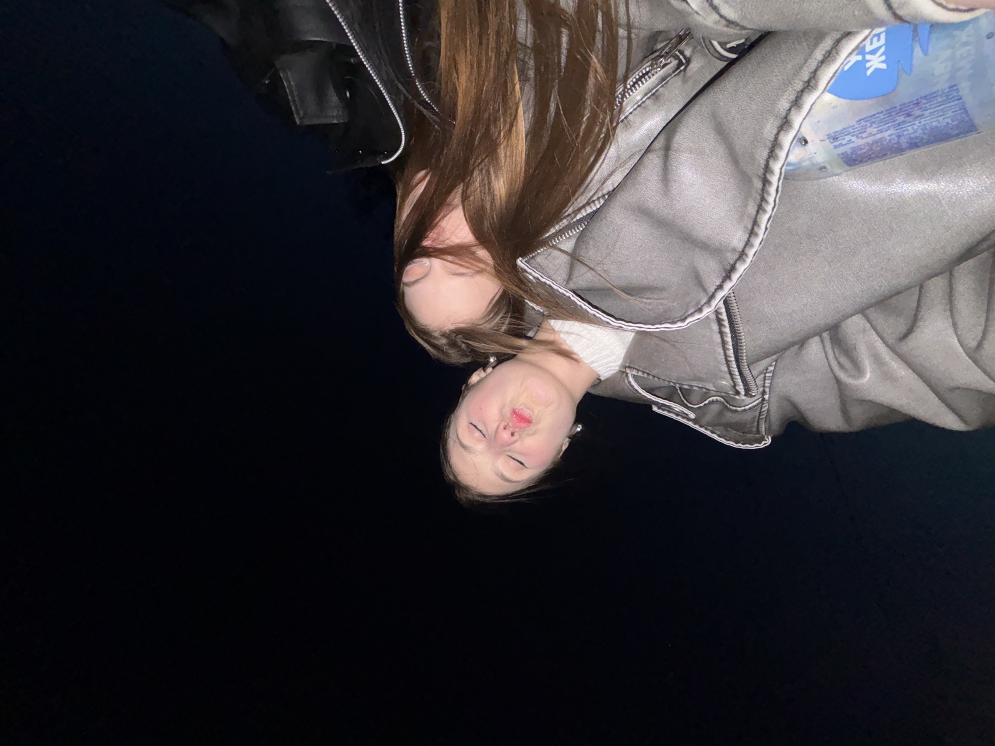 A young woman with brown hair is wearing a gray leather jacket and puckering her lips in the city of Фоки, Russia. There is a water bottle in her jacket. It is nighttime.