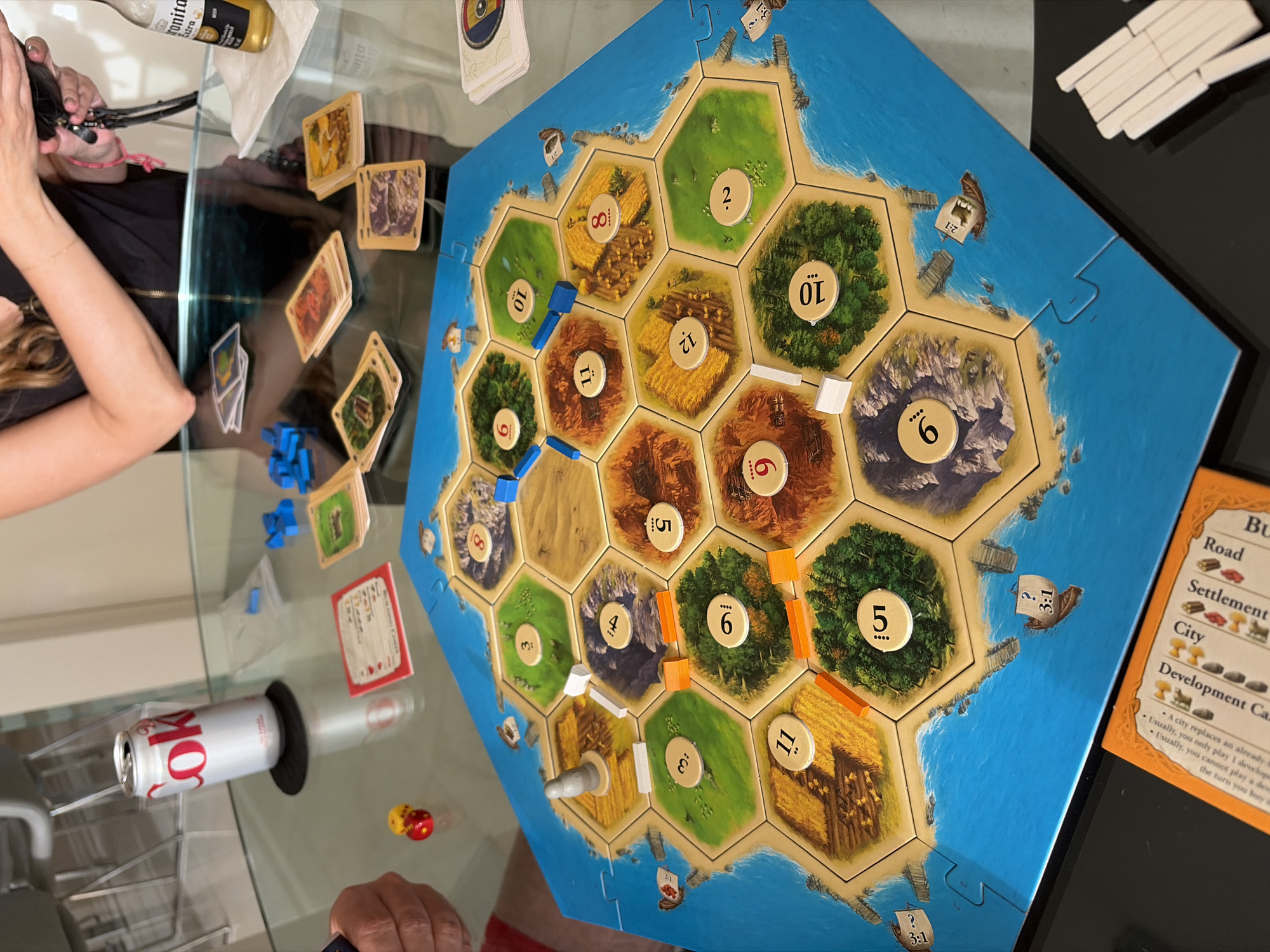 The image depicts a game of Settlers of Catan in progress, likely at a residence. Visible components include the game board with hexagonal resource tiles, number tokens, roads, settlements, and at least one city. Also visible are development cards and resource cards. A red Coke can, Corona beer bottle, and dice are visible. People can be seen at either side of the board.