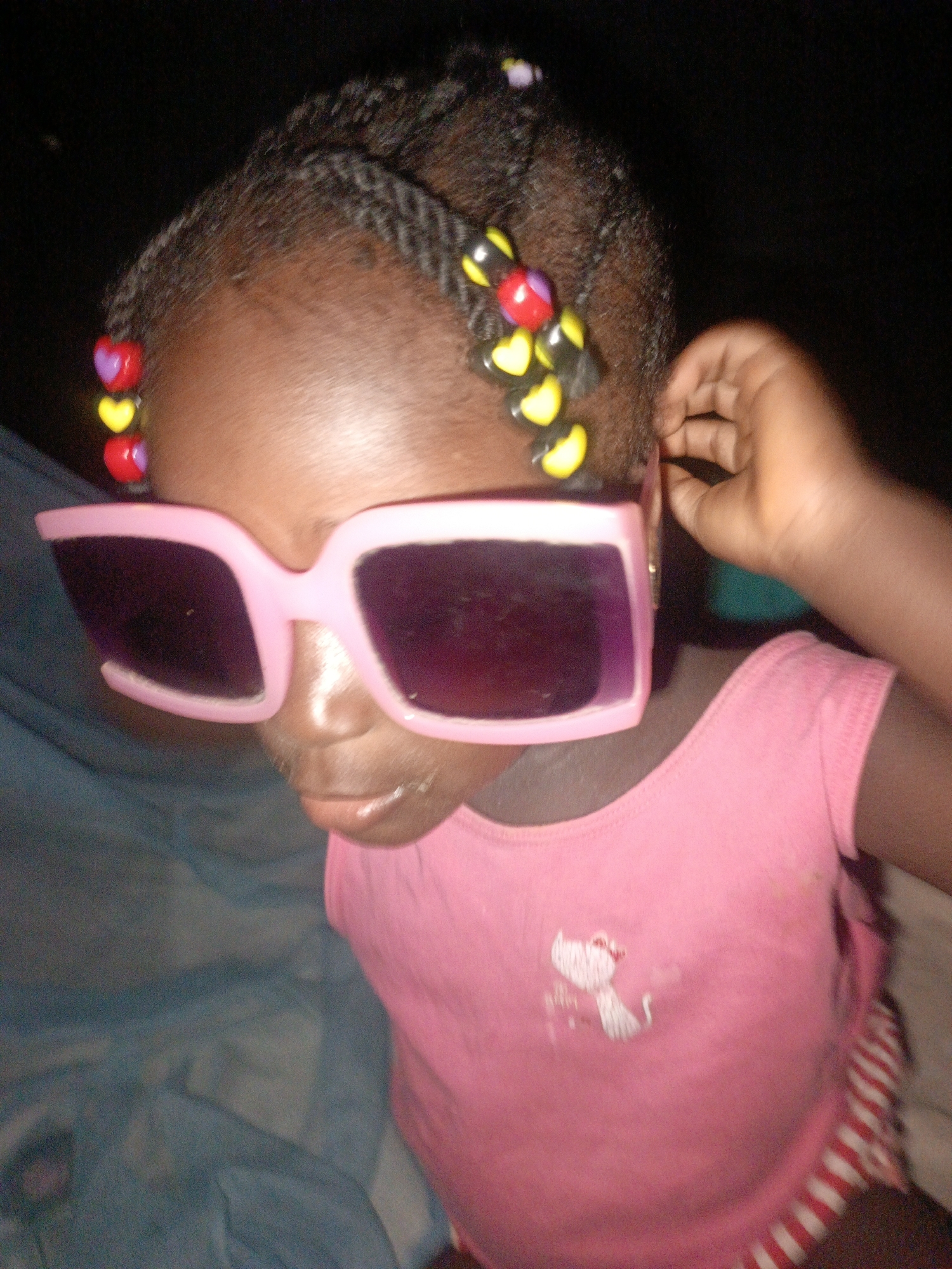 A young Black female child is depicted wearing oversized pink plastic sunglasses and a pink tank top. Her hair is braided with colorful beads, including red, yellow, and purple heart-shaped beads and black circular beads. The child's right hand is visible near her ear. The background consists of dark, out-of-focus elements. The image has a grainy, low-resolution quality.